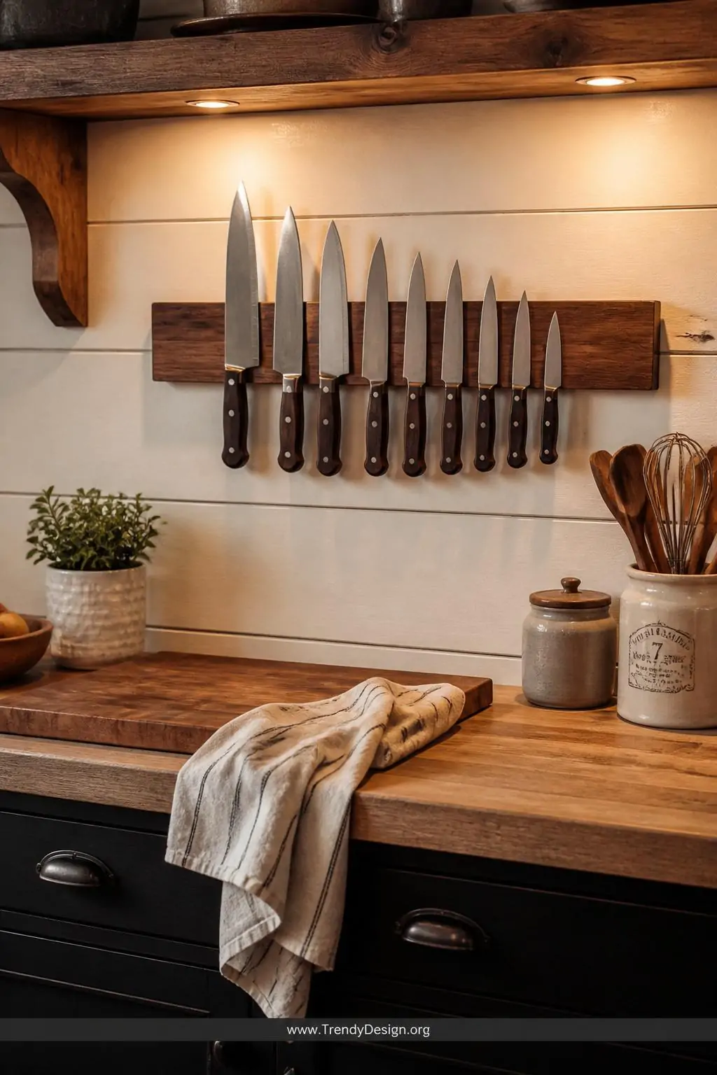 Walnut magnetic knife strip mounted above a butcher block prep counter in a farmhouse kitchen, warm amber overhead lighting casting soft shadows on the wood grain, chef's knives arranged by size against the rich brown strip, medium shot showing the prep zone below with a linen dish towel draped nearby, photorealistic interior design.