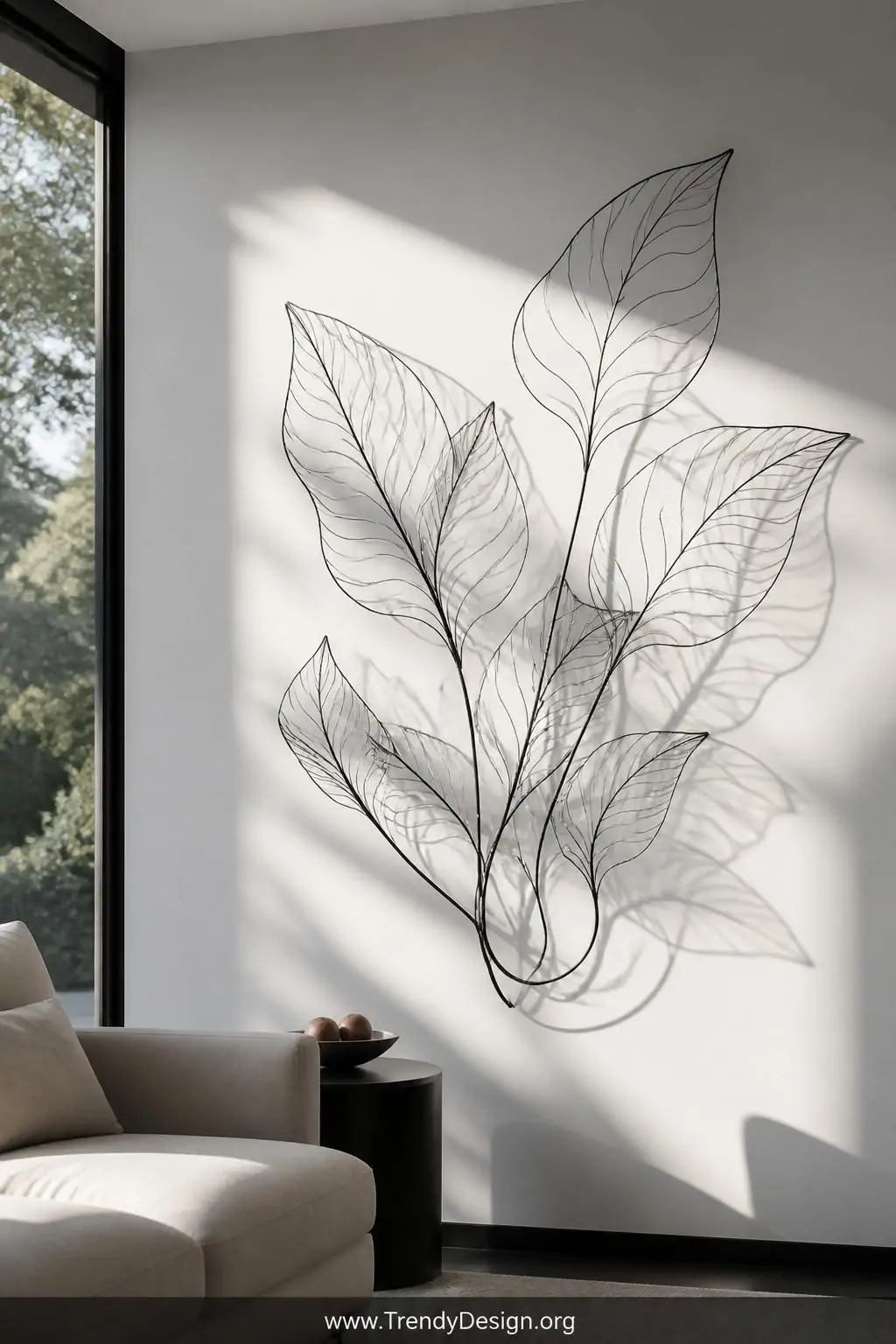 Abstract wire wall sculpture with twisted botanical leaf shapes mounted on a matte white wall, black spray-painted aluminum wire casting dramatic shifting shadows across the surface, modern living room, natural afternoon sunlight streaming from the left, medium shot showing the full sculptural piece against a clean minimalist backdrop.
