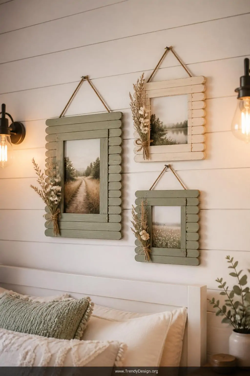 Cluster of three rustic popsicle stick picture frames in varying sizes painted in muted sage green and cream with twine hangers and dried flower accents, arranged on a white shiplap bedroom gallery wall with warm Edison bulb sconces casting golden light across the textured wood grain surfaces, medium shot.