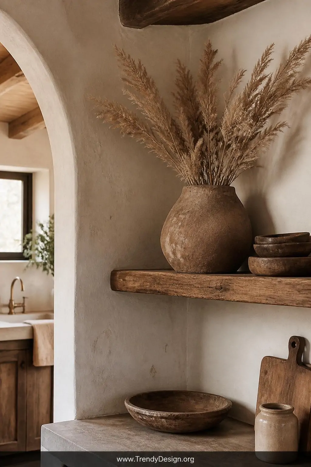 Lopsided pinch pot clay vase with rough textured exterior holding dried pampas grass on a worn wooden kitchen shelf, warm amber light, neutral terracotta tones against whitewashed plaster walls, medium shot of rustic Tuscan-Scandinavian kitchen corner, photorealistic interior design, no people