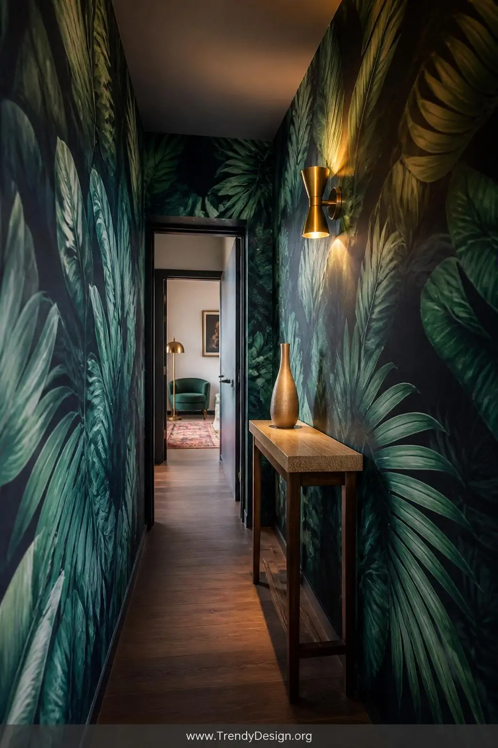 Oversized tropical leaf botanical wallpaper in a narrow hallway, maximalist style — rich deep teal and moody navy palm frond pattern covers floor-to-ceiling walls flanking a slim natural wood console table topped with a single sculptural vase, warm brass wall sconce casting golden upward light, wide shot showing the full dramatic length of the corridor.