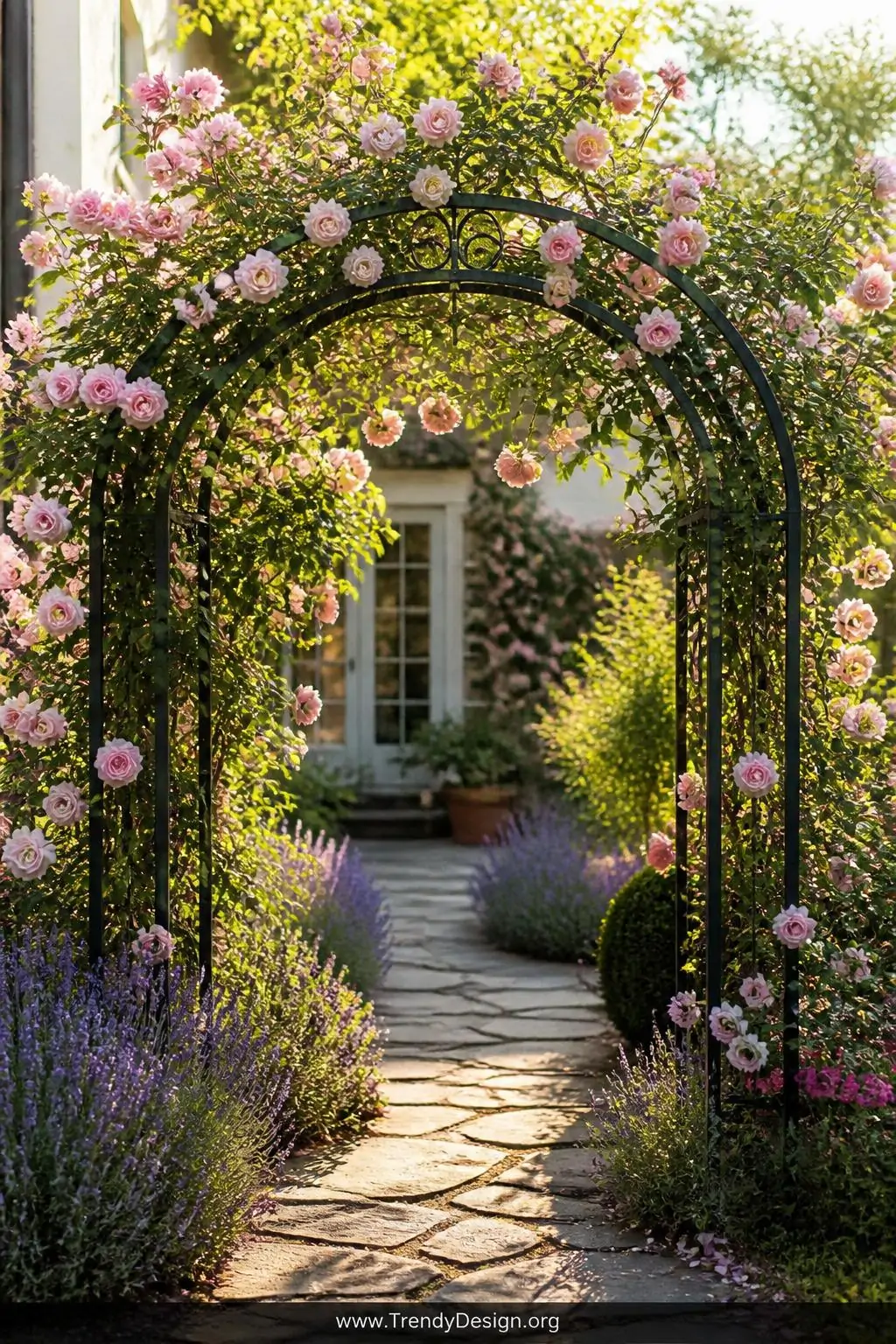 Weathered wrought iron rose arch draped in cascading blush pink 'New Dawn' climbing roses at a cottage garden entrance, flagstone path beneath, purple lavender underplanting at the base, soft golden afternoon light filtering through petals, wide shot, photorealistic interior design photography.