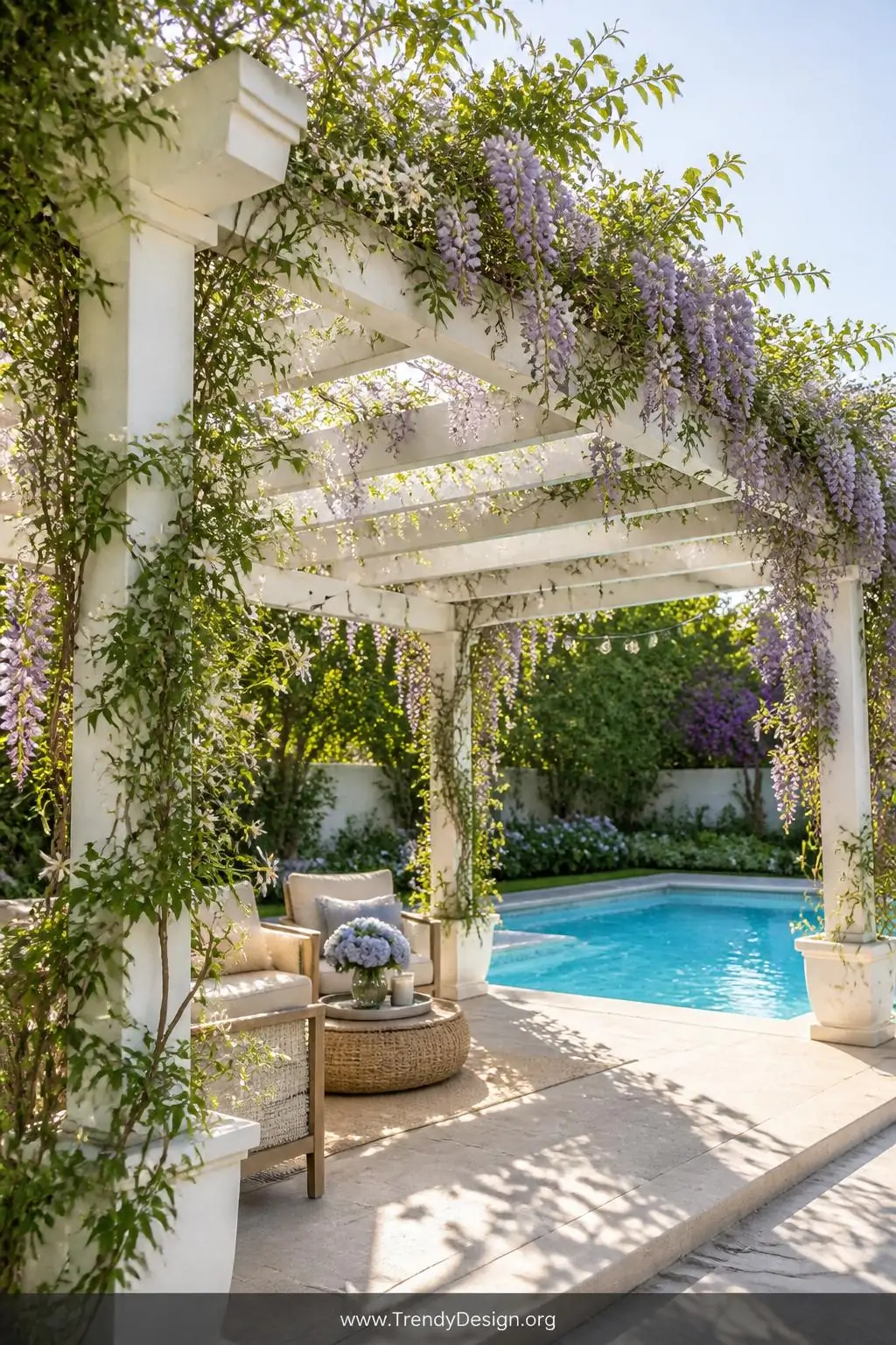 White vinyl pergola draped in cascading wisteria and star jasmine vines over a sunlit pool patio, warm afternoon light bouncing off the bright white frame, lush green tendrils weaving through the lattice roof, string lights threaded along the beams, wide shot showing the full structure against a blue swimming pool.
