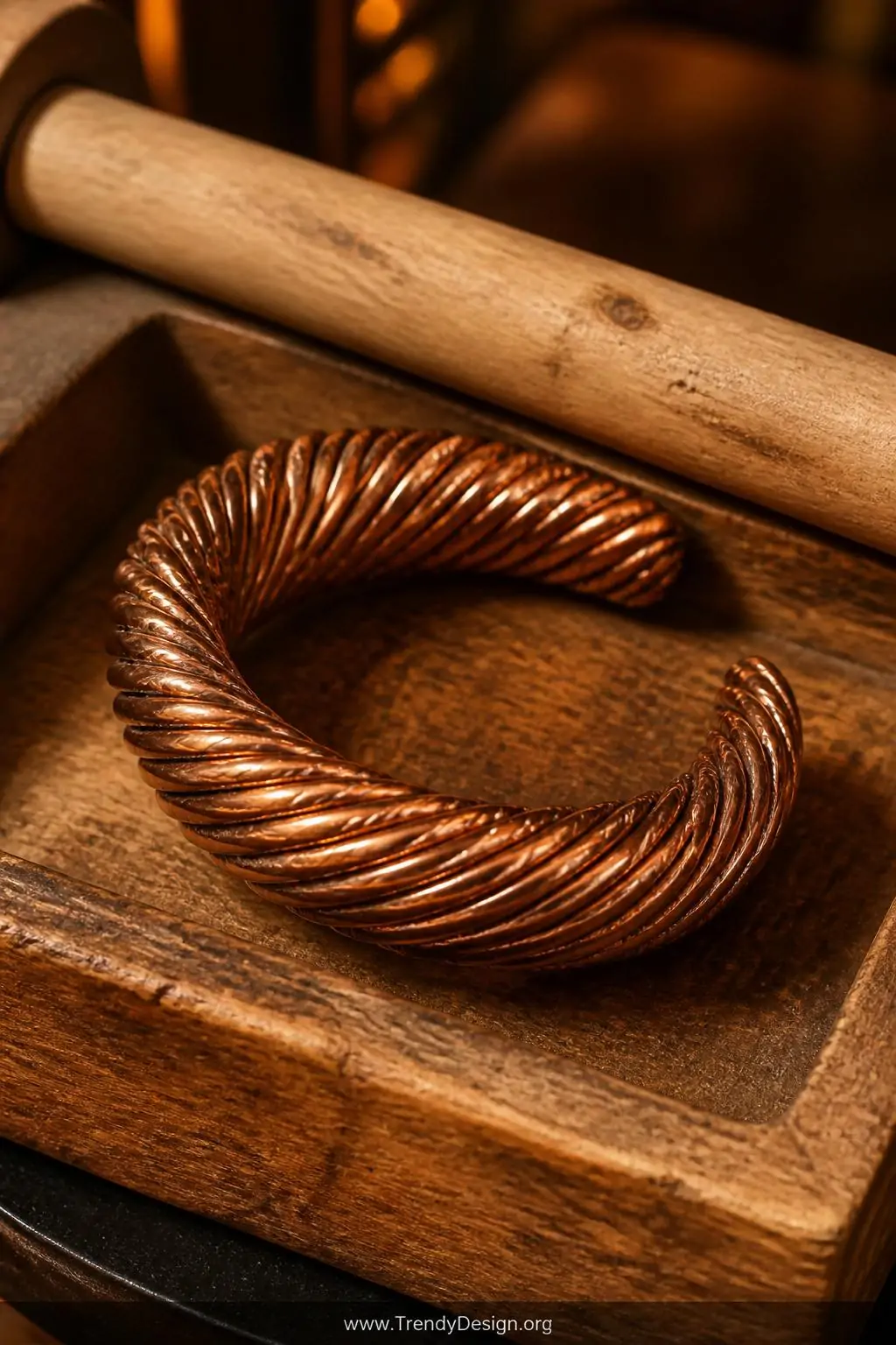 Copper coil bracelet resting on a raw wood jewelry tray beside a dowel rod, warm amber workshop lighting, rich orange-brown patina texture with spiraling grooves catching golden light, closeup