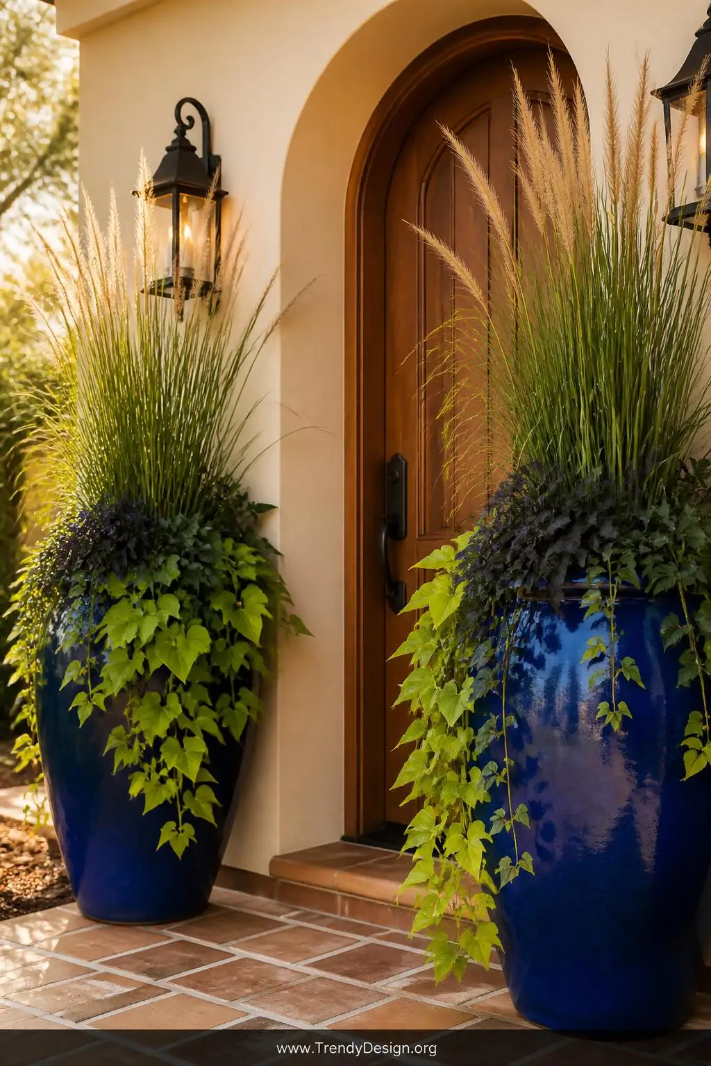 Pair of oversized cobalt blue ceramic urns flanking a wooden front door, wide shot, filled with tall ornamental grass and cascading sweet potato vine in deep green and chartreuse, rich terracotta pathway underfoot, warm golden afternoon light illuminating the bold glazed surfaces, photorealistic exterior entryway design.