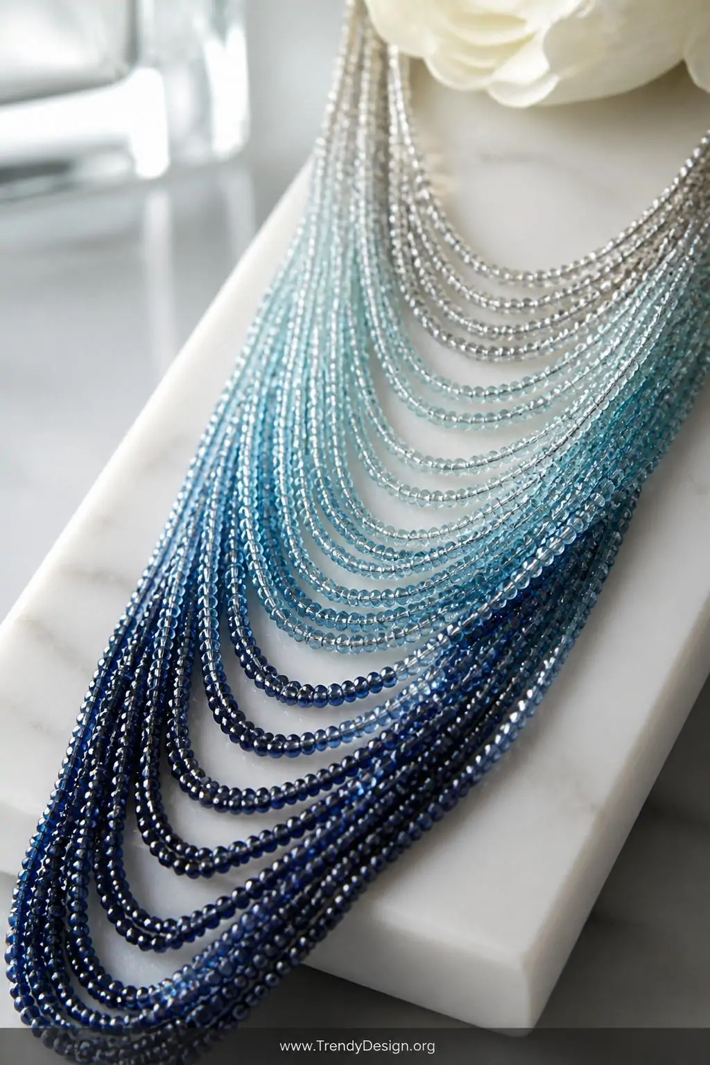 Tiny seed bead strands in cascading ombre blues and silvers arranged in graduating waterfall lengths on a white marble vanity tray, soft diffused window light, extreme closeup showing shimmer and delicate drape.