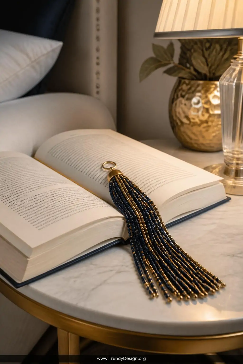 Seed bead tassel bookmark draped over an open hardcover book on a marble side table, navy and gold tiny beads cascading in 20 lush strands with a polished gold jump ring at the top, soft warm reading lamp glow highlighting the metallic shimmer, elegant bedroom nook, closeup shot.
