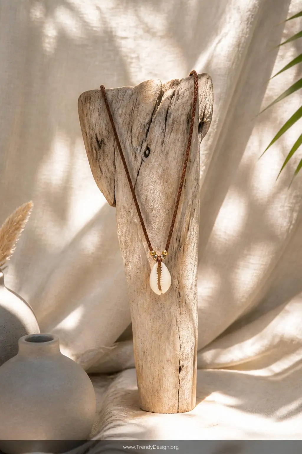Single cowrie shell necklace on braided brown leather cord displayed draped over a weathered driftwood jewelry stand, sun-bleached linen backdrop, gold bead accents catching warm afternoon light, medium shot, photorealistic interior design