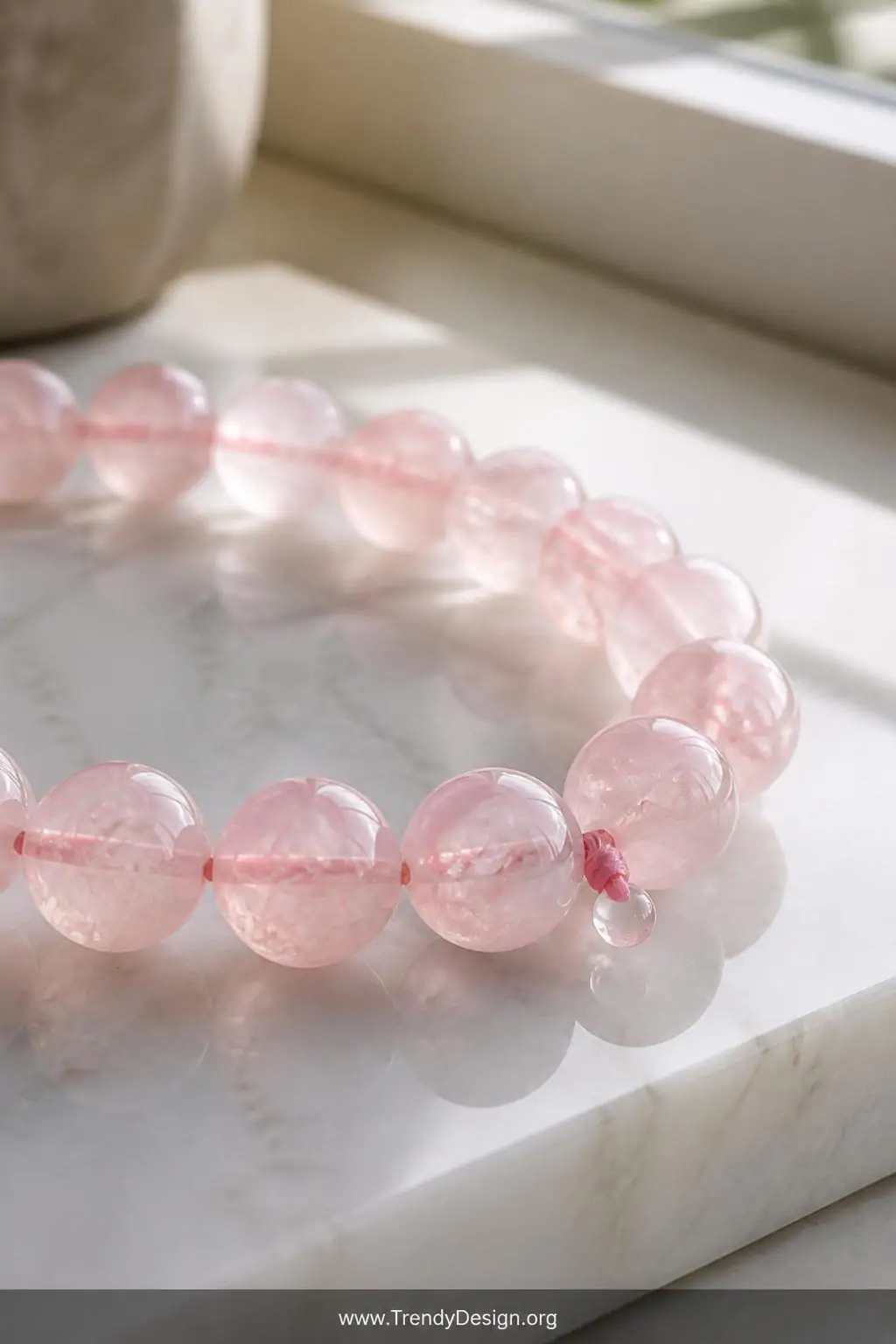 Strand of uniform 8mm rose quartz beads on a white marble surface, soft natural daylight streaming from a nearby window, macro closeup showing the translucent blush-pink stones with their delicate internal veining and silk-tied elastic knot sealed with a glossy drop of glue.