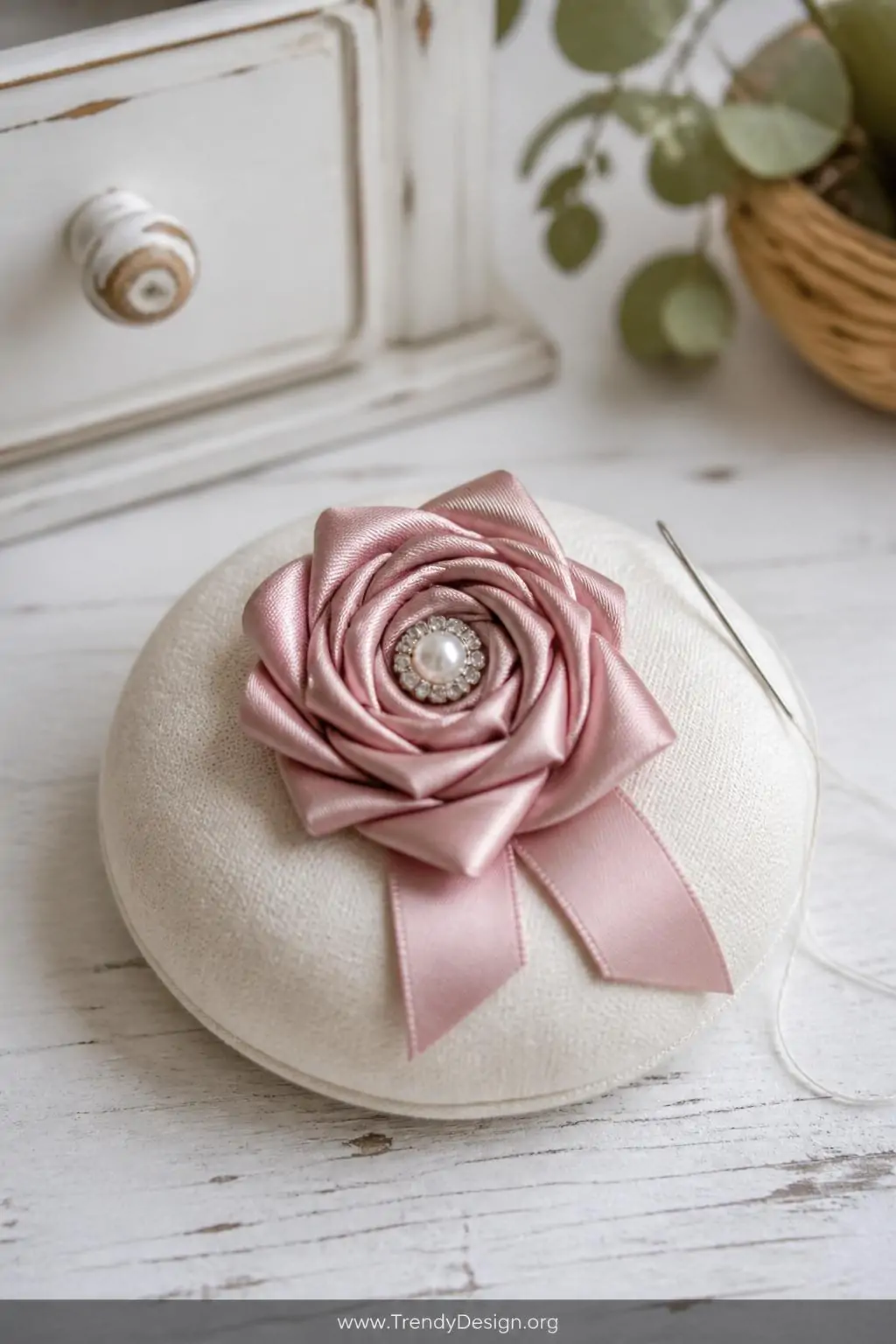 Satin ribbon rosette brooch on a cream linen pin cushion beside a needle and thread on a weathered white farmhouse craft table, soft diffused window light, closeup