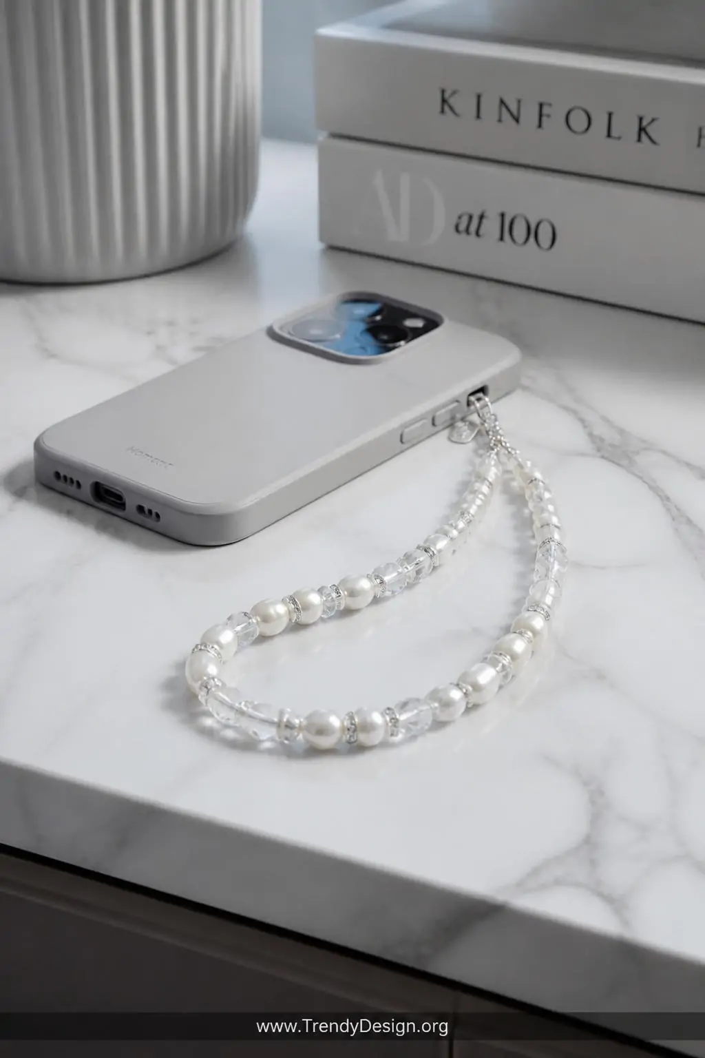 Faux pearl and crystal beaded phone charm draped over a sleek white marble desk surface beside a minimalist phone case, soft cool studio lighting, closeup