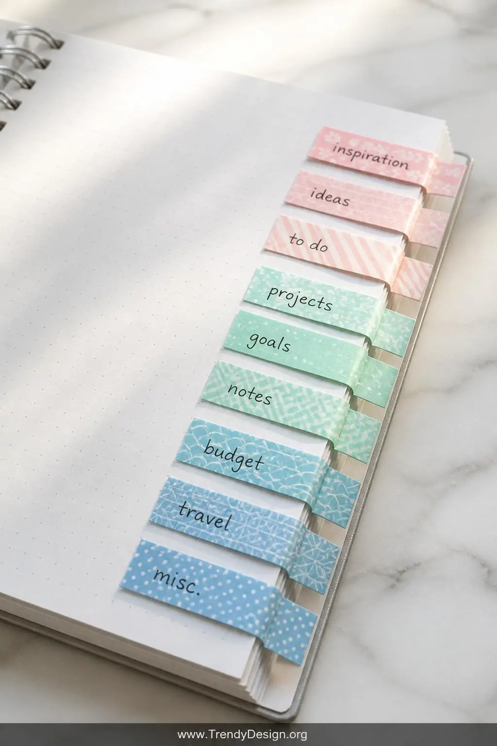 A closeup shot of a open spiral notebook lying flat on a white marble desk, with colorful washi tape tabs folded over page ed