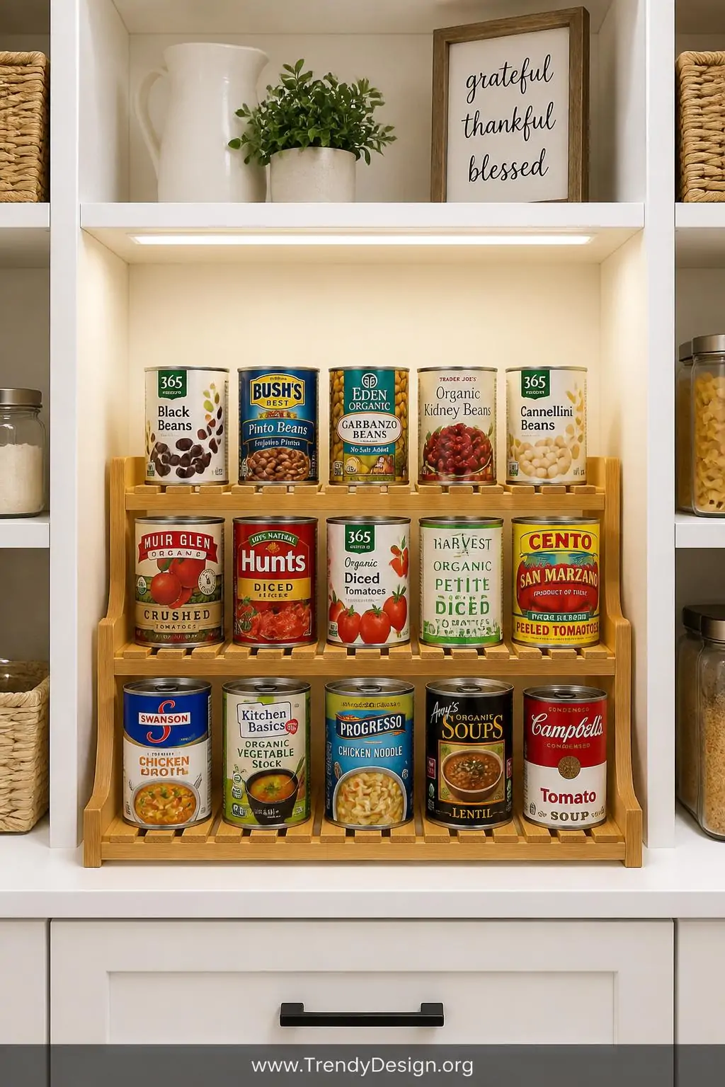 Bamboo tiered shelf organizer inside a bright white pantry, modern farmhouse style, loaded with colorful canned goods labels all facing forward across three rising stadium-style rows, warm overhead LED lighting casting soft shadows on the slatted wood surface, medium shot showing full shelf width with clean painted wood shelving on either side.
