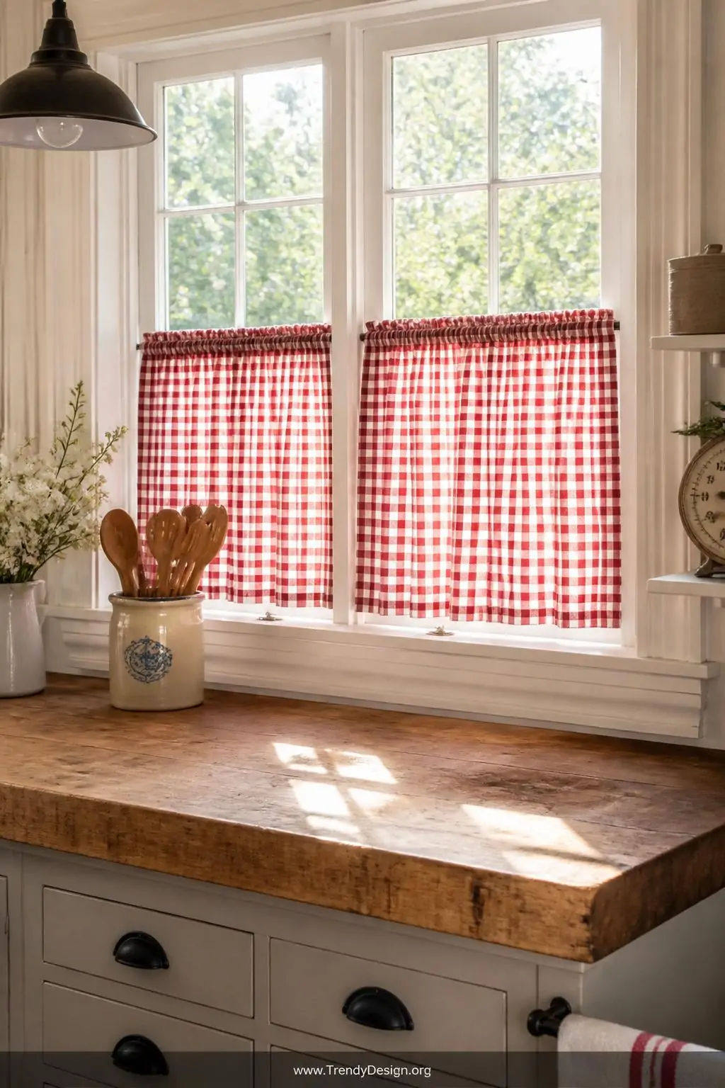 Red and white gingham café curtains hung at the lower half of a bright cottage kitchen window, medium shot, cheerful daylight streaming in above the panels onto a worn butcher block counter with a ceramic crock of wooden spoons nearby, photorealistic interior design.