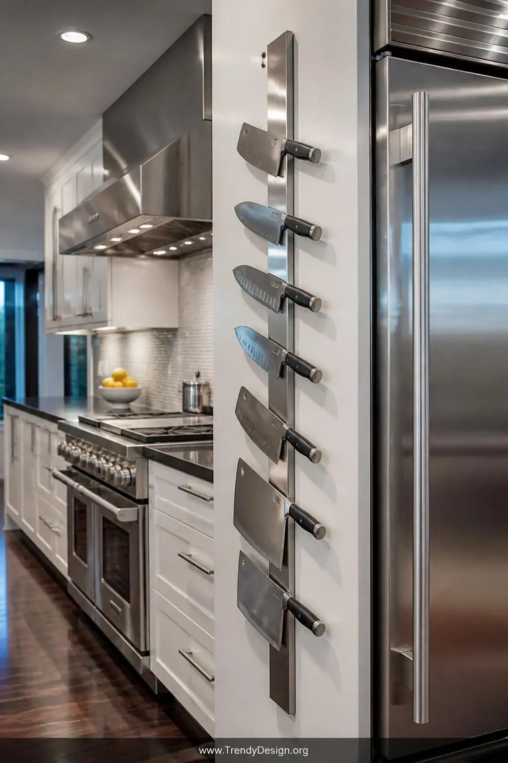 Brushed stainless steel magnetic knife strip mounted vertically on the side of a white cabinet in a sleek contemporary kitchen, cool recessed lighting reflecting off polished metal surfaces, heavy cleavers and chef's knives held firmly in a vertical column beside matching stainless appliances, medium-wide shot capturing the industrial-modern aesthetic, photorealistic interior design.