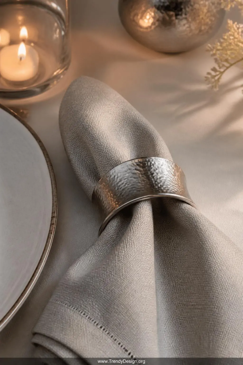 Hammered silver cuff bracelet sliding around a folded linen napkin on a white dinner table, elegant sculptural tablescape, candlelight casting soft shadows across the engraved metallic surface, closeup.