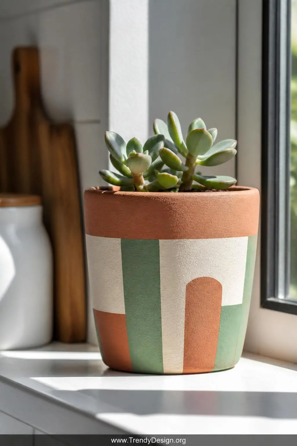 Hand-painted terracotta flower pot with bold geometric stripes in terracotta orange, cream, and sage green holding a small succulent on a sunlit kitchen windowsill, natural daylight streaming across rough clay texture, closeup