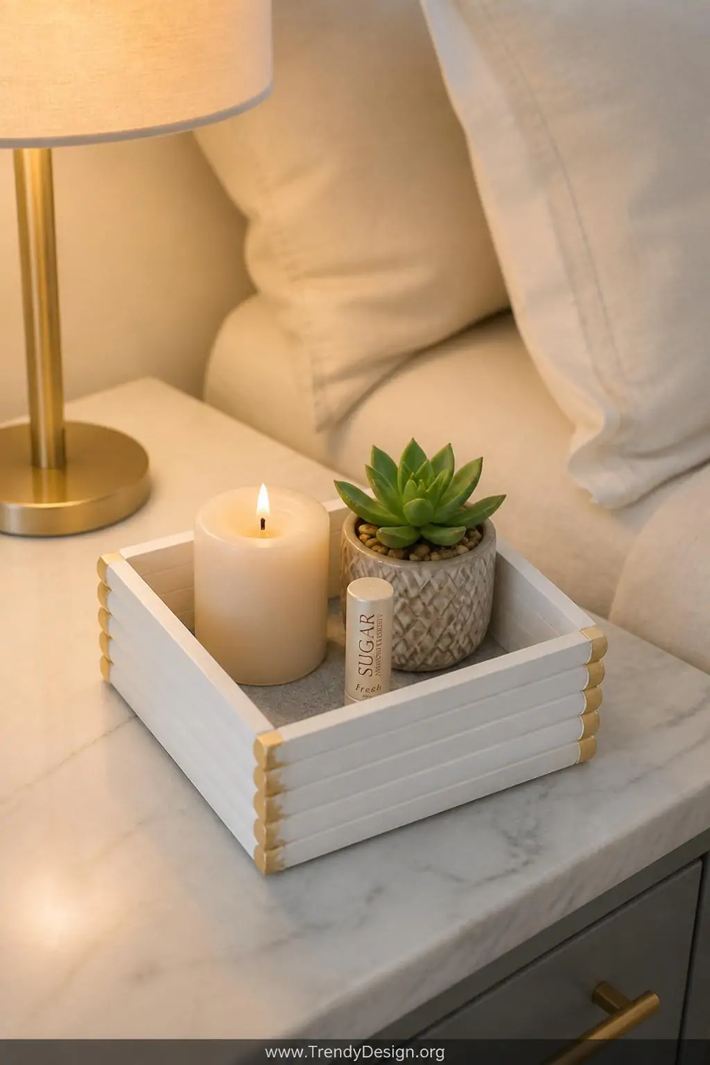 Small square popsicle stick organizer tray painted in matte white with gold-trimmed edges holding a lit cream pillar candle, lip balm, and a tiny succulent on a white marble nightstand beside soft linen bedding, warm bedside lamp glow illuminating the felt-lined base, closeup shot.