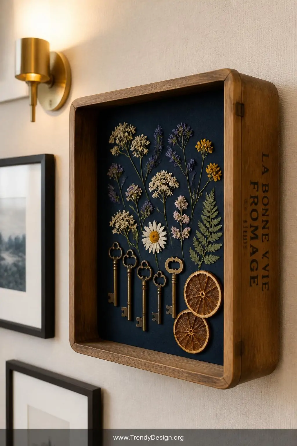 Mounted wooden cheese box shadow box on a cream linen-textured wall displaying pressed wildflowers, vintage brass keys, and dried citrus slices against a deep navy painted interior, gallery wall style living room, soft ambient sconce lighting, closeup shot