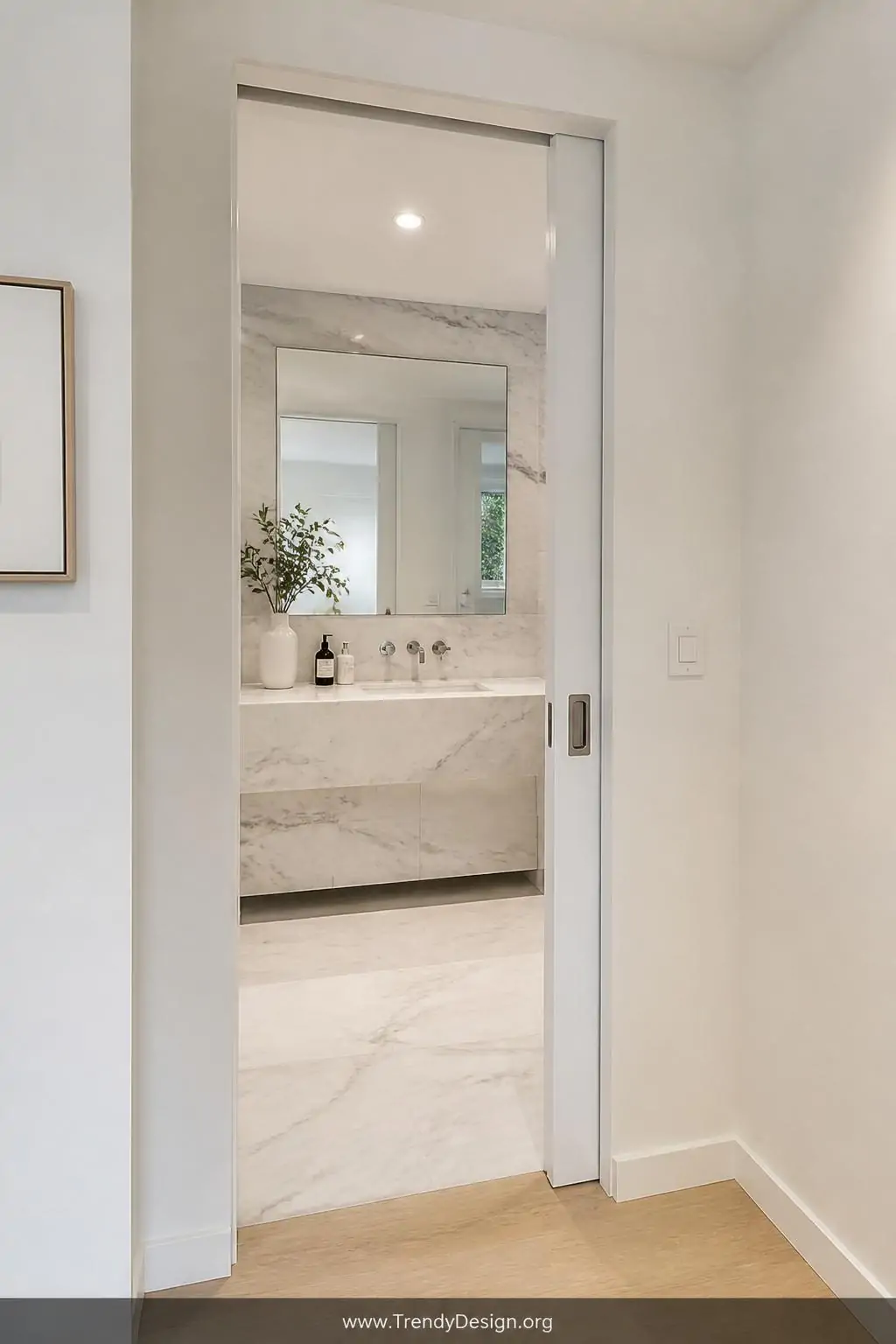 Seamless pocket door flush inside a white drywall cavity of a compact ensuite bathroom, medium shot, recessed brushed nickel pull handle barely visible, soft overhead LED lighting, crisp white walls meeting pale oak flooring, door completely vanished into the wall revealing a marble vanity beyond, photorealistic interior design.