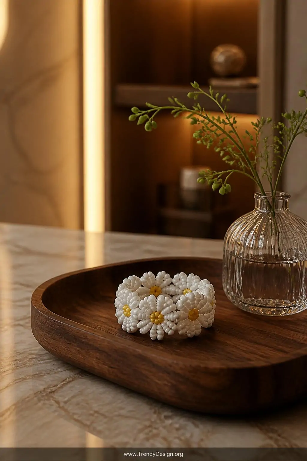 Three-dimensional white floral seed bead ring with raised daisy petals and a yellow center bead resting on a dark walnut jewelry tray beside a small glass vase, warm golden ambient light, medium shot capturing dimensional depth and petal layering