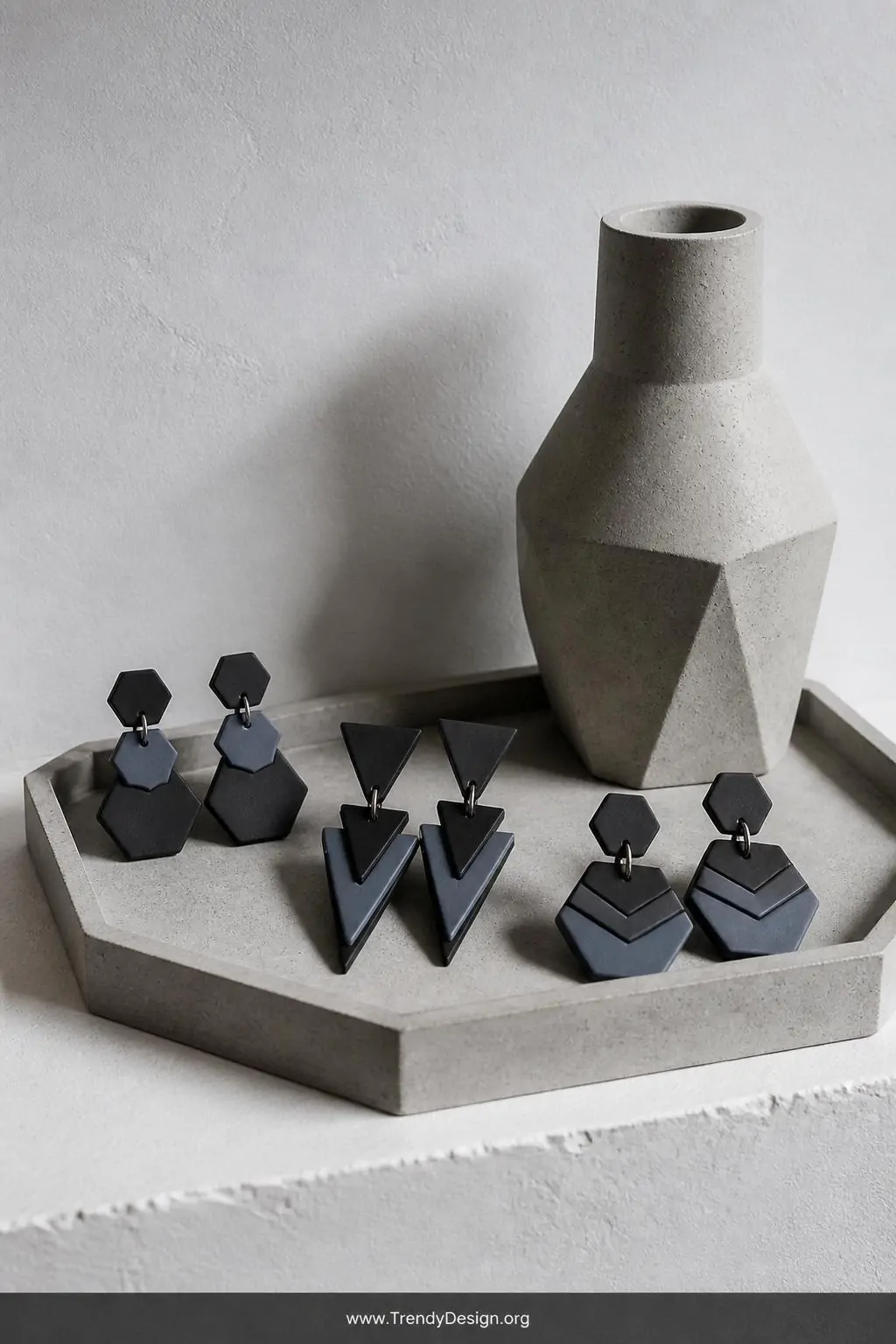 Geometric hexagon and triangle polymer clay earrings in matte charcoal and dusty slate two-tone layers arranged on a minimalist concrete tray beside a single architectural vase on a white plaster shelf, cool directional studio lighting emphasizing sharp angular edges and editorial matte finish, medium shot.