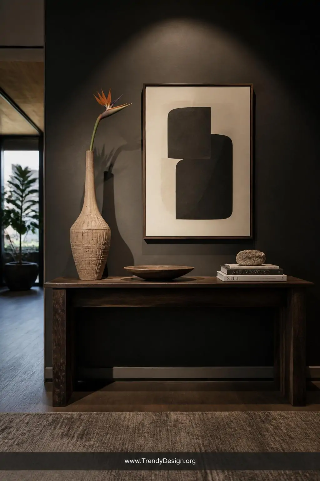 Tall sculptural clay vase with carved geometric patterns and elongated neck displaying a single bird of paradise stem on a dark wood entryway console, dramatic directional spotlight casting sharp shadows, boutique-style modern interior, wide shot showing full console styling against a moody charcoal accent wall, photorealistic interior design, no people