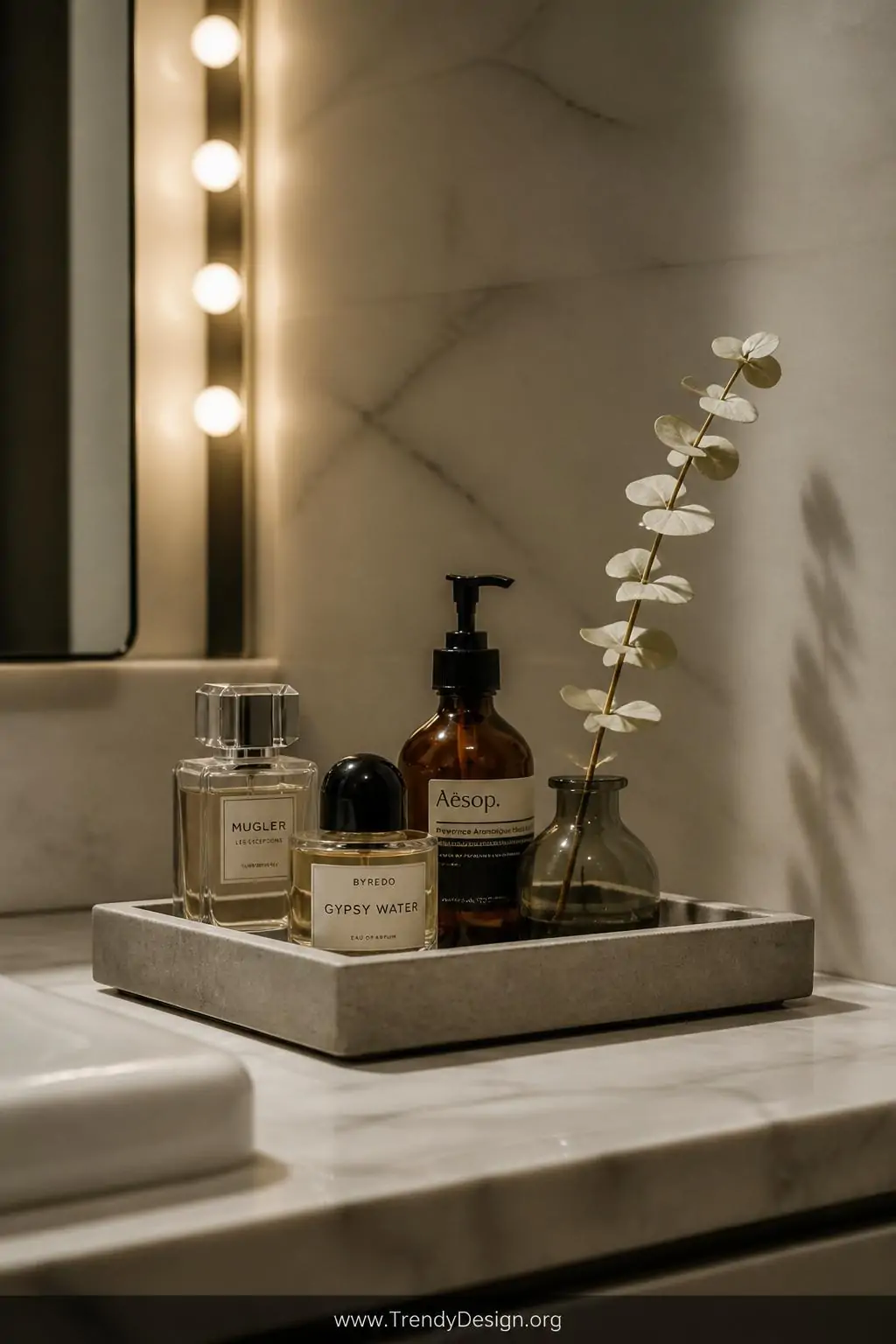 Shallow concrete tray on a marble bathroom vanity in a spa-style bathroom, holding glass perfume bottles, a amber-toned lotion dispenser, and a small bud vase with a single white eucalyptus sprig, soft warm vanity bulb lighting casting gentle shadows, closeup shot.