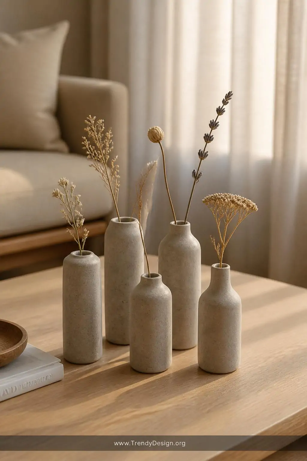 Cluster of five mini concrete bud vases in varying heights grouped on a light oak coffee table, each holding a single dried flower stem, soft afternoon light filtering through linen curtains, warm neutral tones, closeup shot, photorealistic interior design, no people.