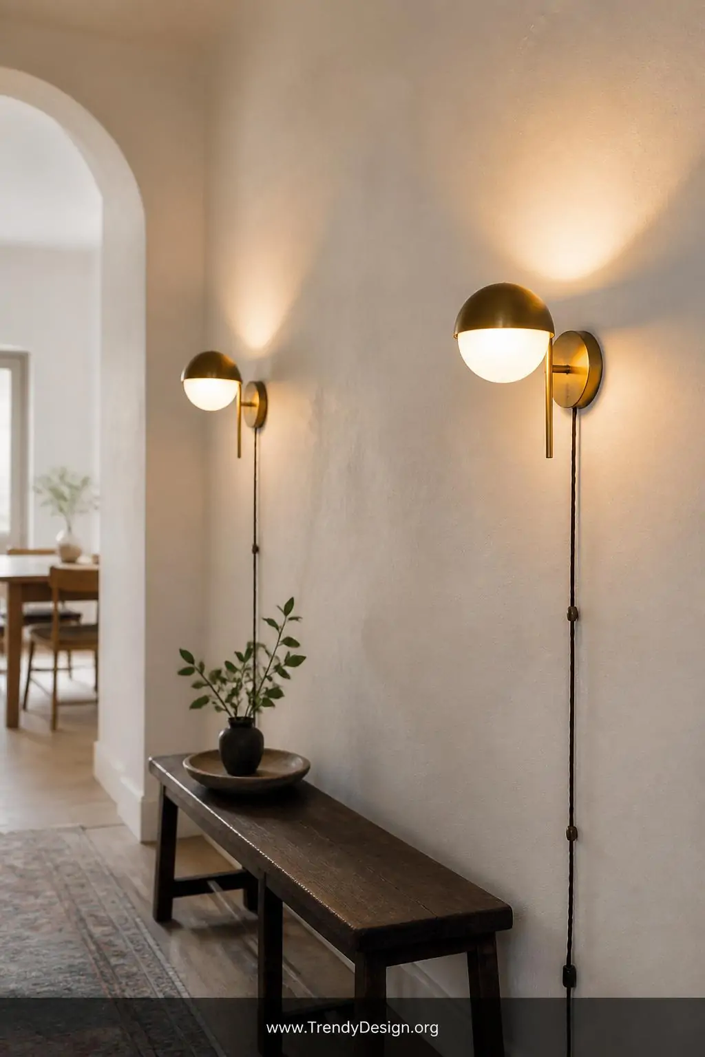 Pair of aged brass globe wall sconces mounted on a soft white plaster hallway wall, fabric-corded plug-in style casting warm upward and outward light across subtle textured wallpaper, medium shot, photorealistic interior design.