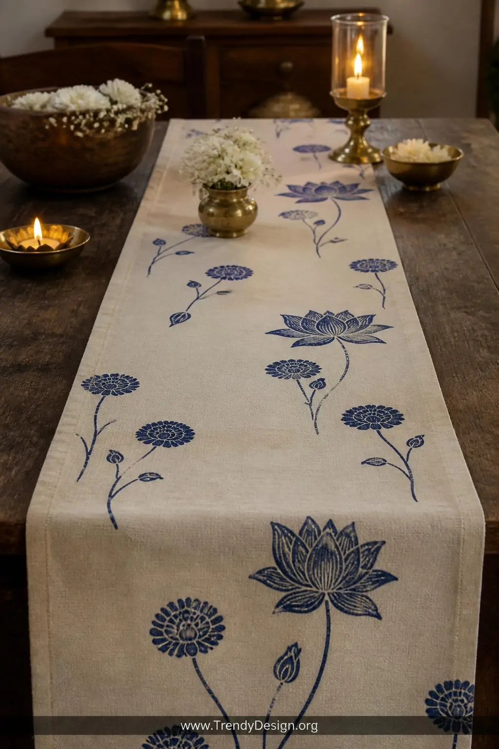 Cotton table runner with block-printed indigo lotus and marigold floral motifs centered on a rustic dining table, scattered blooms in deep indigo on cream cotton, warm candlelight, dark walnut table surface, closeup shot showing organic flower spacing and rich pigment absorption