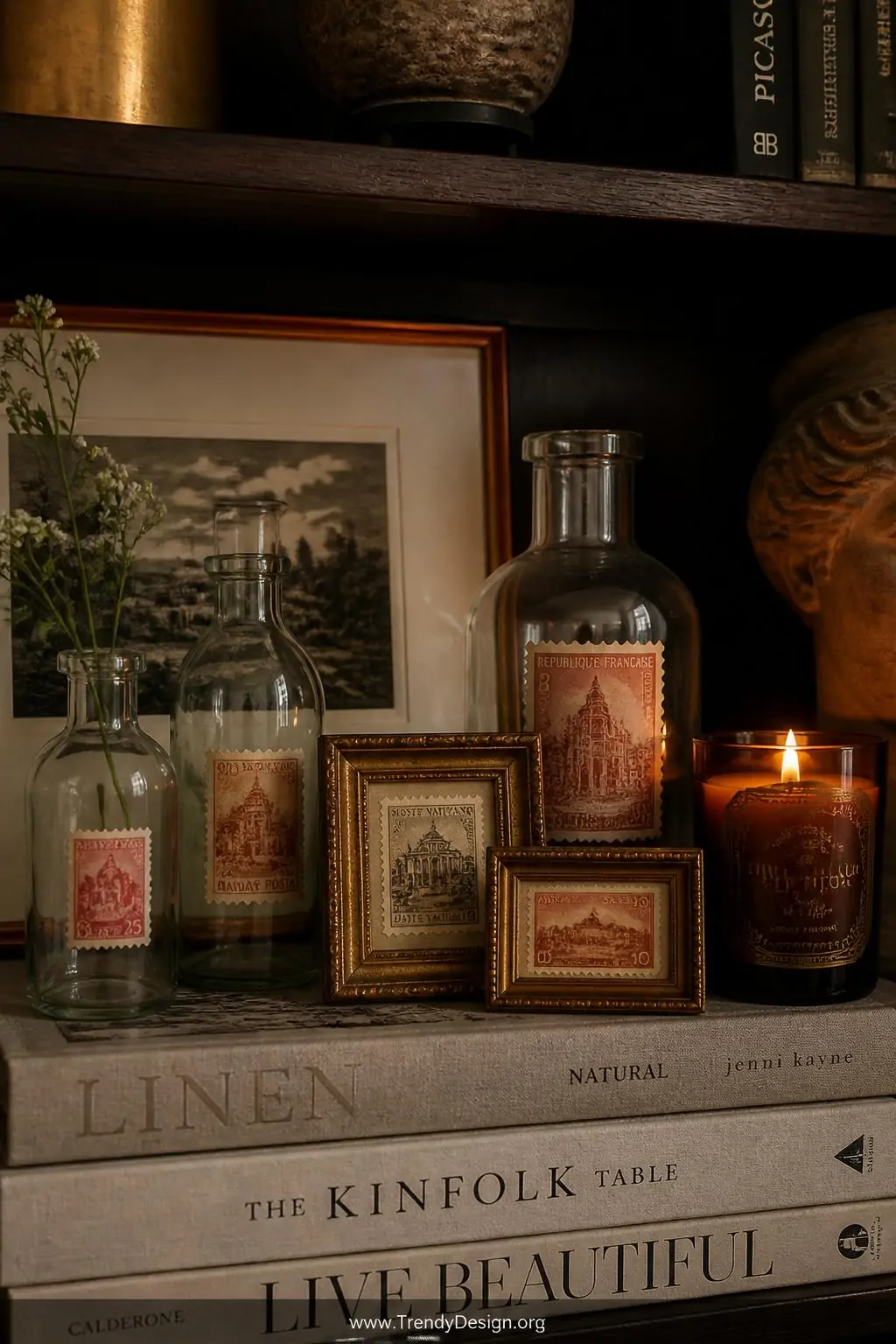 Sepia and faded red vintage postage stamp stickers applied to small gilded frames and plain glass vases on a dark wood bookshelf, warm amber candlelight, closeup of an eclectic styled shelfie with stacked linen-covered coffee table books.