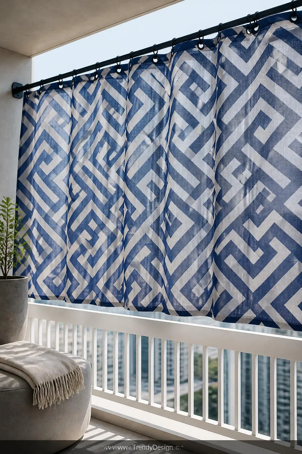 Outdoor fabric privacy panel in a bold navy geometric print hanging from matte black grommets on a tension rod, modern urban balcony, contemporary style, breathable mesh weave billowing softly in natural light, crisp white railing beneath, a sleek concrete planter and folded linen throw nearby, bright midday diffused light, wide shot.