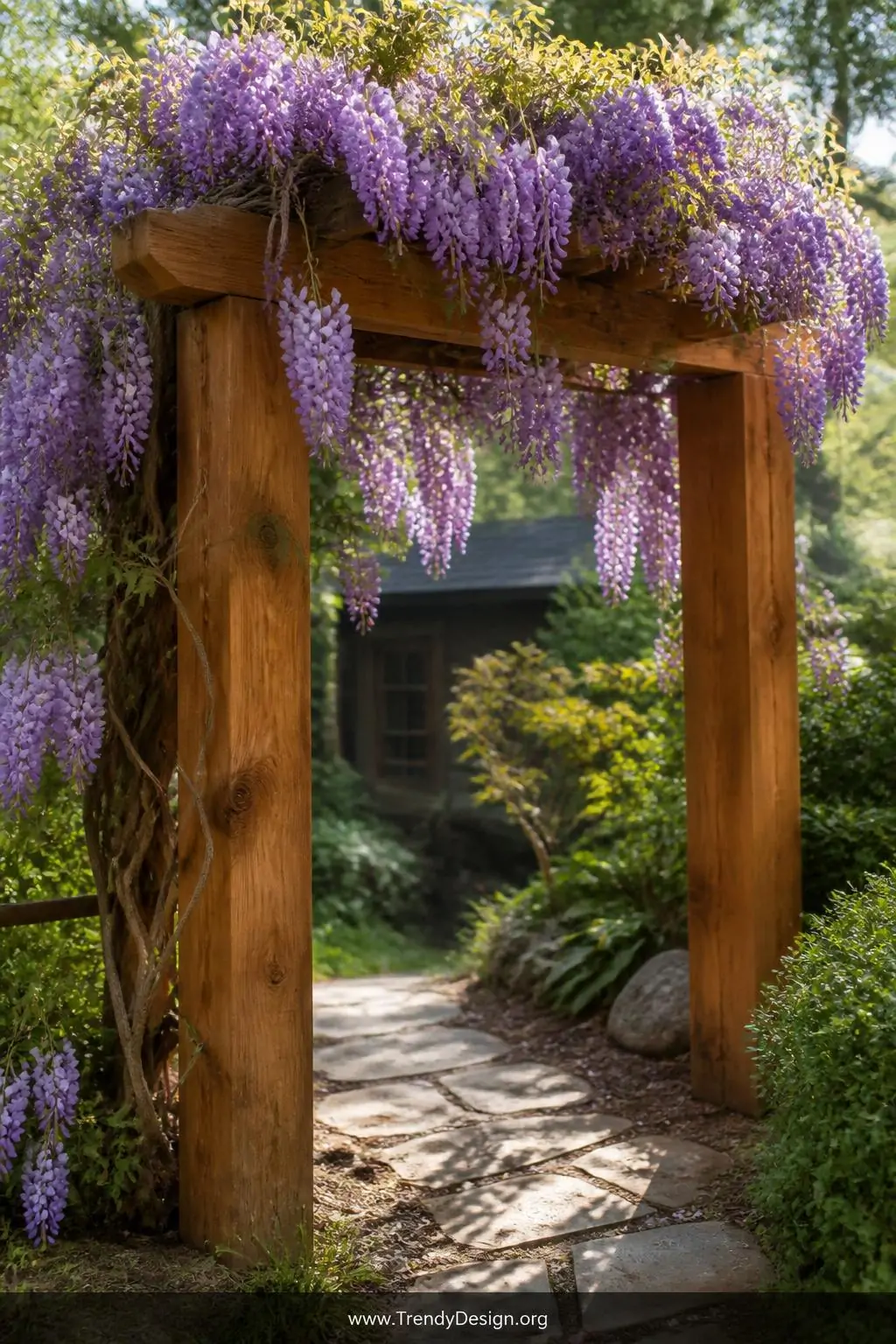 Chunky cedar wooden garden arch in a naturalistic backyard, rustic organic style, raw honey-stained timber posts anchored deep into earth supporting heavy cascading purple wisteria blooms with pendulous grape-like flower clusters, rough-hewn wood grain texture visible against the violet softness overhead, dappled afternoon light filtering through the dense floral canopy, medium shot framing the arch from a slight angle.