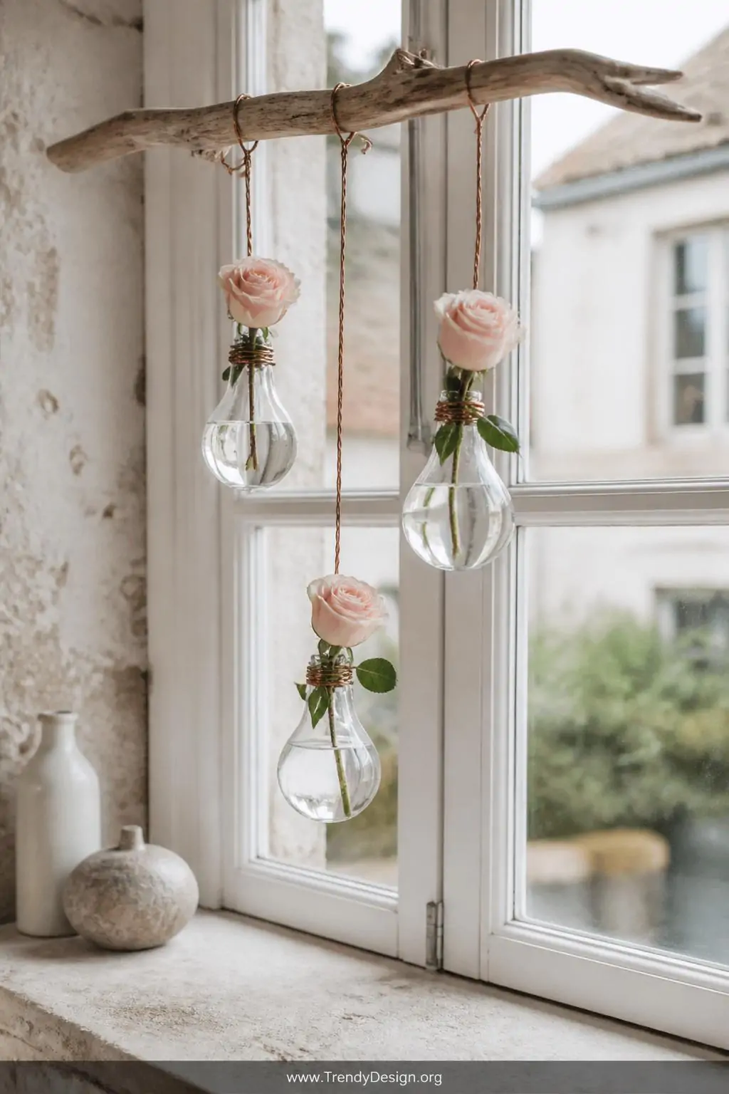 Hollowed clear glass light bulb vase holding a single blush rose stem, suspended by twisted copper wire from a weathered driftwood branch mounted on a whitewashed French cottage windowsill, soft diffused natural daylight, three bulb vases hanging at staggered heights, wide shot.
