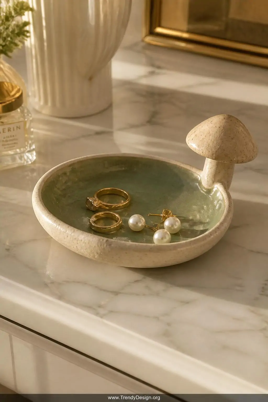 Clay mushroom ring dish with speckled texture and a small sculpted mushroom perched on the rim sitting on a marble vanity dresser top, the dish interior painted dusty sage green holding gold rings and pearl earrings, warm golden hour side lighting, closeup shot, photorealistic interior design