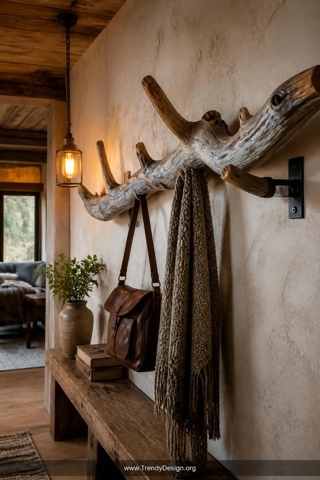 Thick gnarled driftwood branch coat rack mounted with black iron brackets on a rough plaster cabin wall, natural silver-grey and honey-brown tones with visible knots and organic curves, leather satchel and chunky knit scarf draped from natural stub hooks, warm amber pendant light glowing nearby, wide shot, photorealistic interior design.