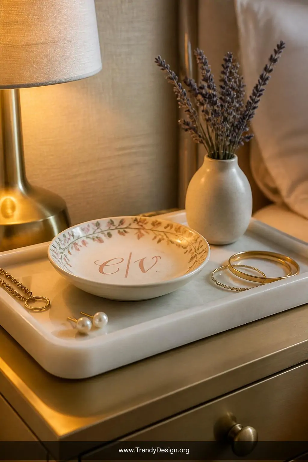 A closeup of a soft ivory marble vanity tray on a brushed brass nightstand holding a hand-painted ceramic jewelry dish with d