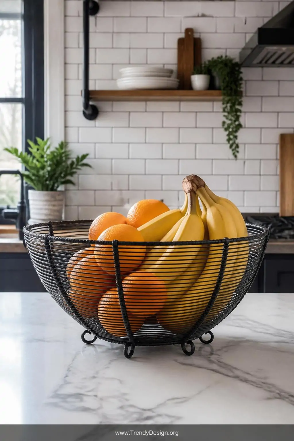 Matte black wire fruit bowl filled with bright oranges and yellow bananas sitting on a white marble kitchen counter, industrial-farmhouse kitchen with subway tile backsplash, natural daylight from a nearby window catching the coiled wire texture, closeup shot emphasizing the open weave structure and colorful fruit contrast.