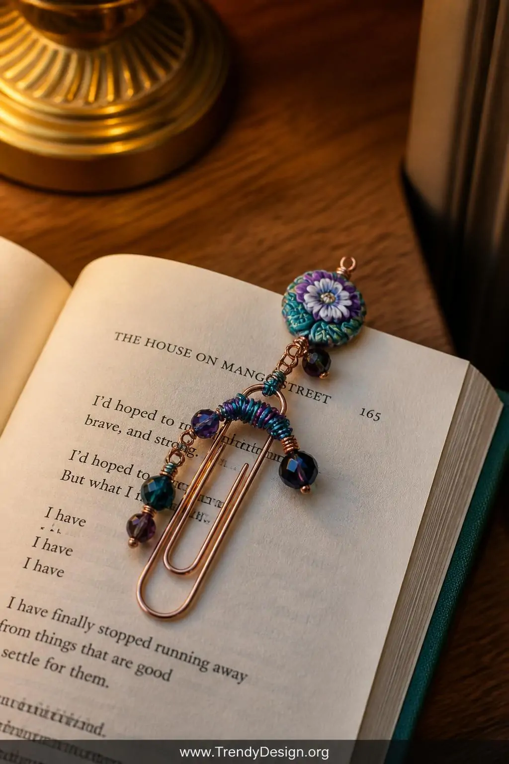 Decorative paperclip bookmark with colorful wire wrapping and tiny bead charms resting across the open pages of a hardcover book on a wooden bedside table, warm golden lamp light illuminating jewel-toned beads and a small floral polymer clay charm peeking over the page edge, closeup shot.