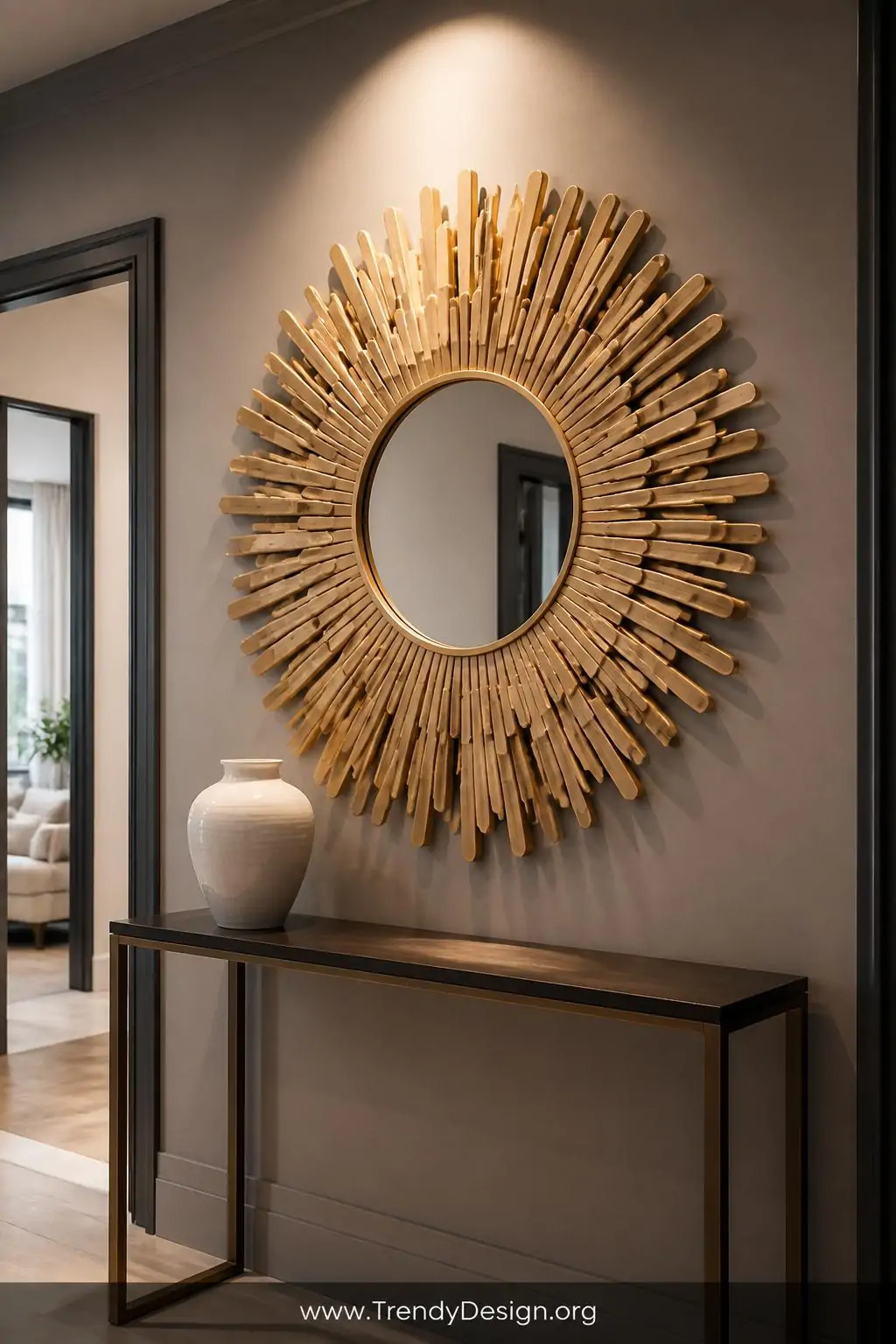 Large sunburst popsicle stick mirror with sticks layered outward in a circular pattern and painted entirely in brushed gold, mounted on a soft gray entryway wall above a slim console table with a white ceramic vase, dramatic warm spotlight highlighting the metallic texture and dimensional depth, wide shot.