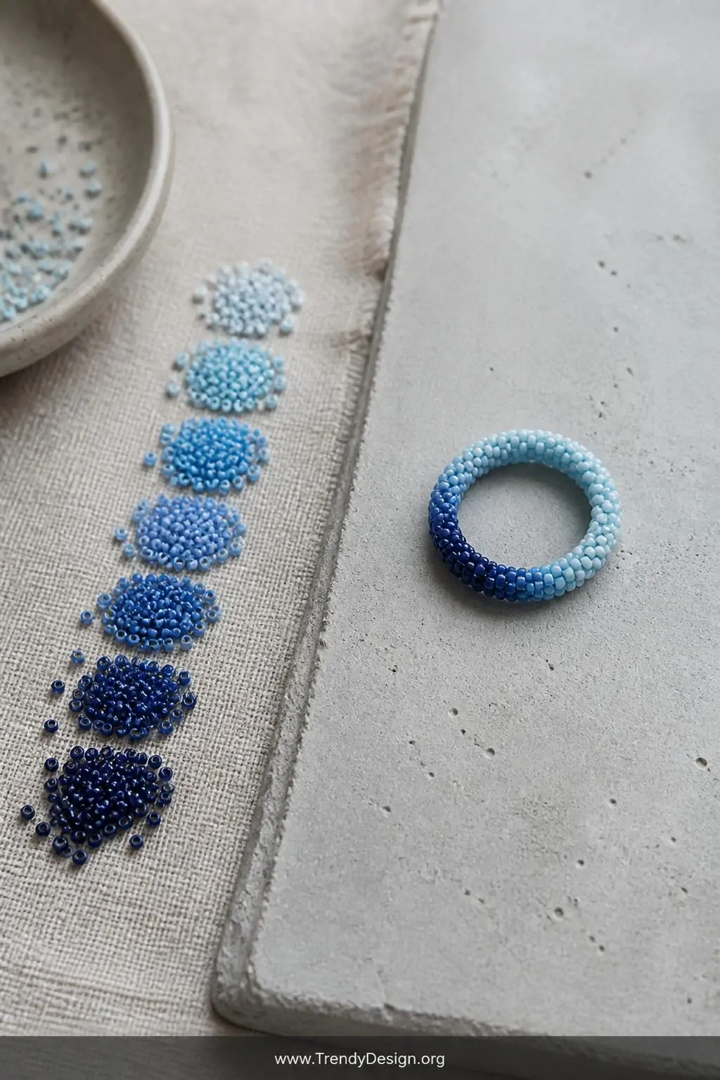 Ombre gradient seed bead ring transitioning from deep navy to pale sky blue displayed on a smooth concrete slab surface beside scattered loose beads arranged in gradient order on a linen beading mat, cool natural daylight, wide styled flatlay shot