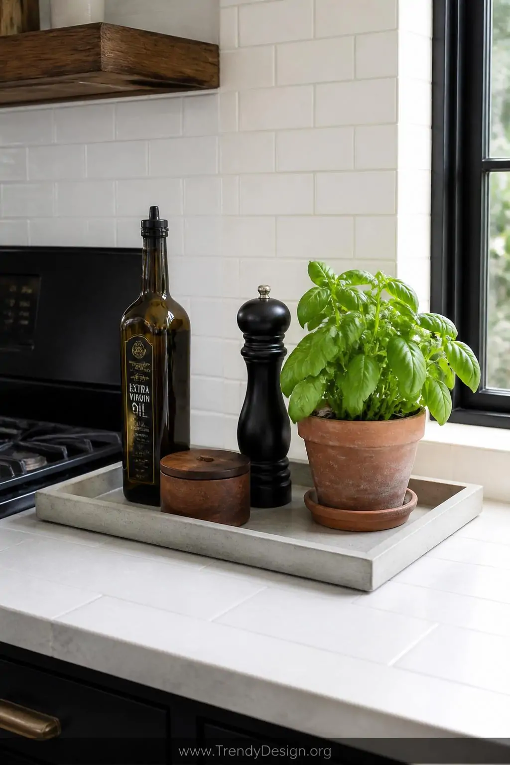 Large concrete tray on a white subway-tiled kitchen counter near a matte black stove, holding a tall amber glass olive oil bottle, a wooden salt cellar, a black pepper grinder, and a small terracotta pot of fresh basil, natural daylight streaming through a nearby window, medium shot.