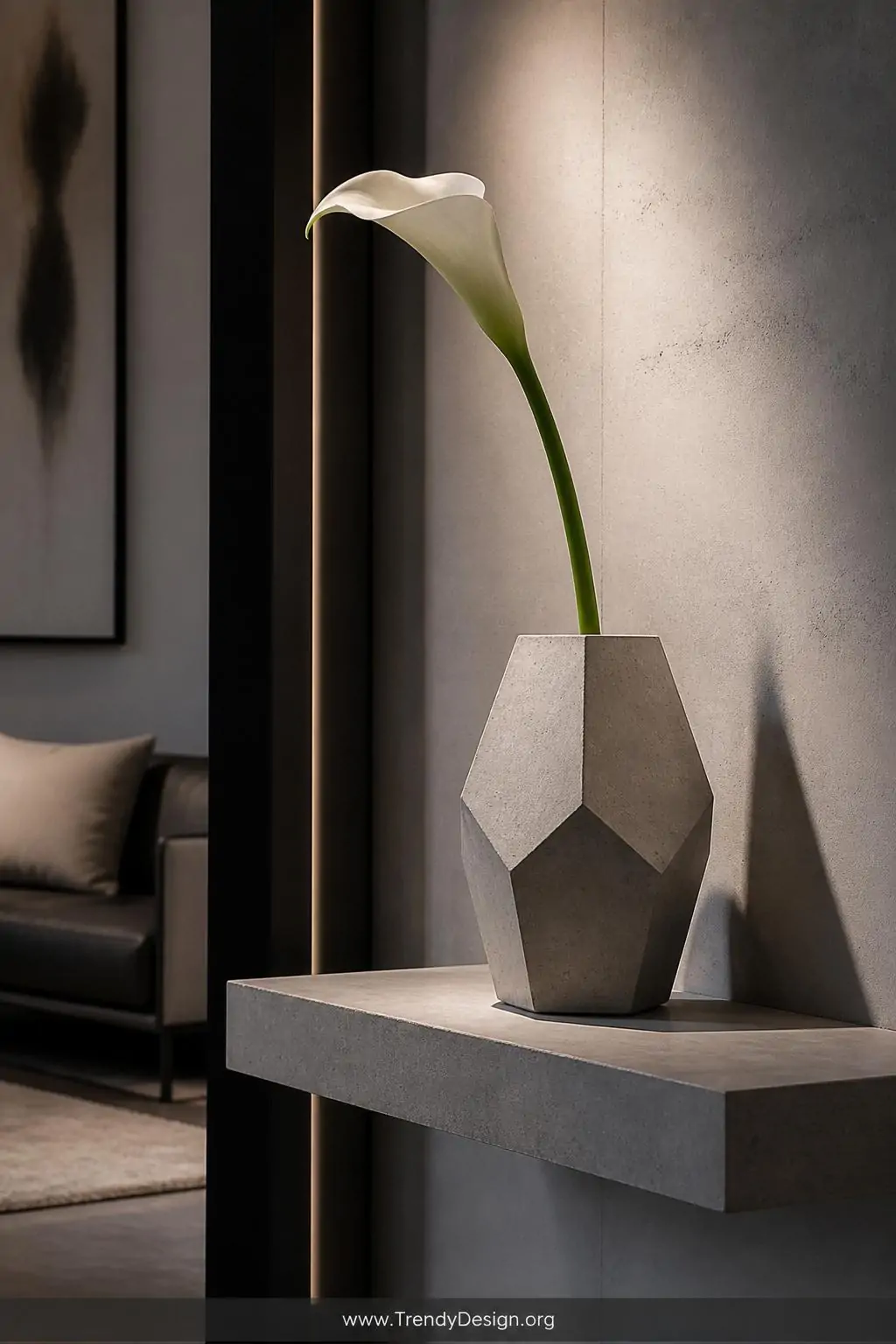 Hexagonal faceted concrete vase with sharp angular edges holding a single structural calla lily on a floating shelf in a modern living room, cool gray and white palette, dramatic directional spotlight, medium shot, photorealistic interior design, no people.