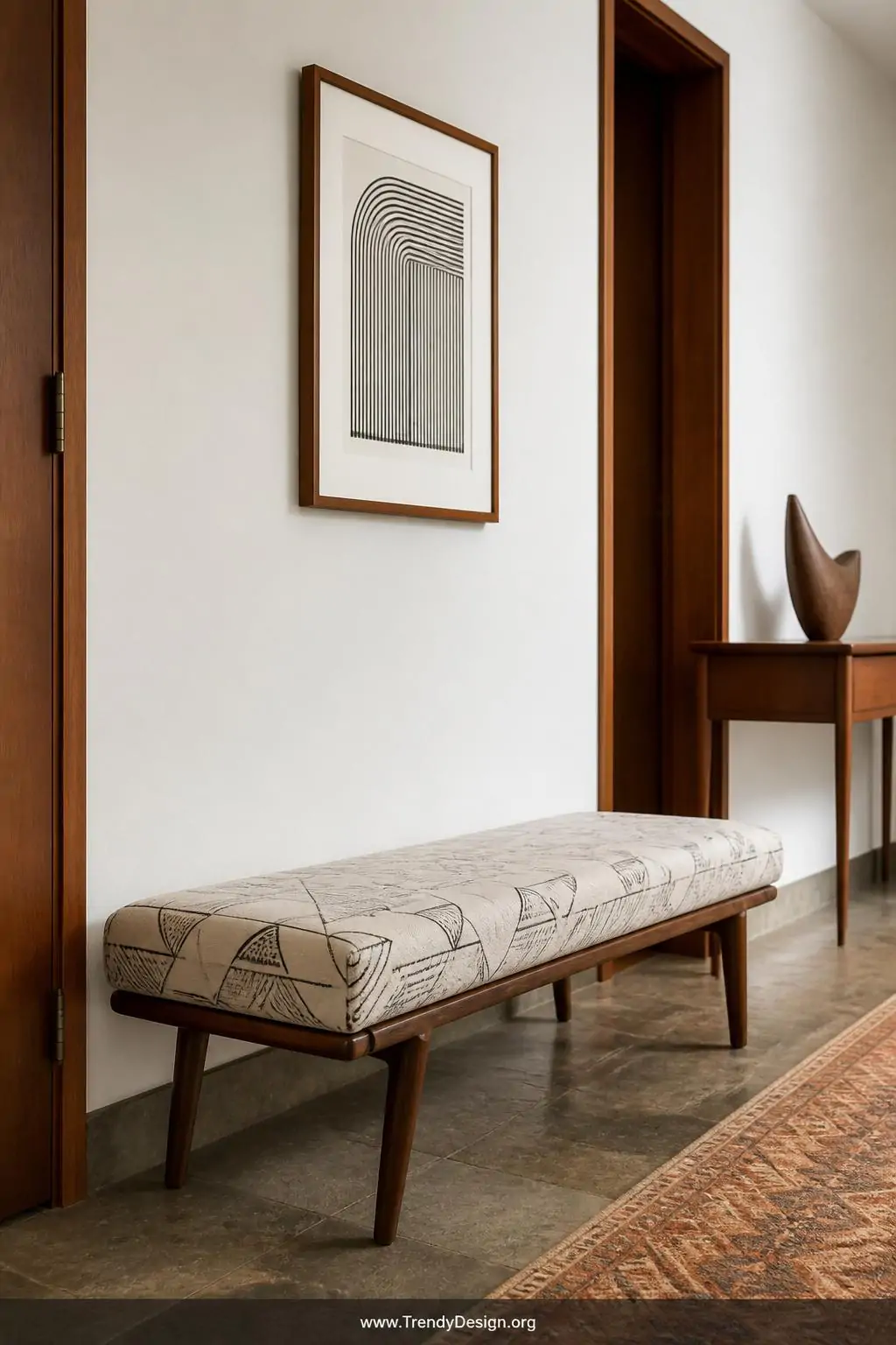 Low-profile mid-century modern bench with tapered walnut legs and clean geometric-patterned linen seat in a narrow entryway, architectural retro style — warm teak tones glowing against a white wall beside a single framed piece of minimalist art, uncluttered negative space and a slim console table with a single sculptural object nearby, medium shot.