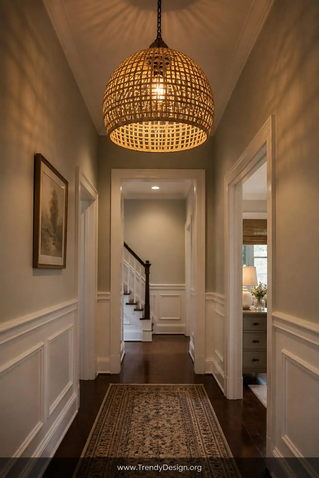Woven rattan pendant light hanging at the entrance of a narrow hallway, open-weave frame spilling warm amber light downward onto a patterned runner rug and white trim woodwork below, medium shot looking upward, photorealistic interior design.