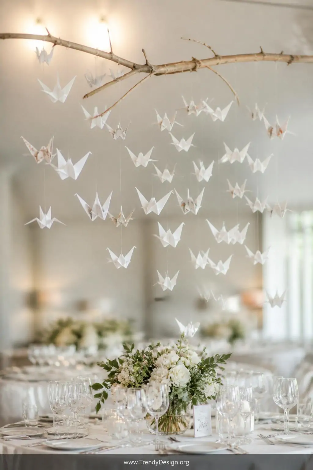 Origami crane mobile of thirty white and patterned paper cranes suspended on invisible fishing line from a natural wood branch above a wedding tablescape, soft diffused overhead light catching each delicate fold, wide dreamy shot showing cranes floating at varying depths over ivory place settings.