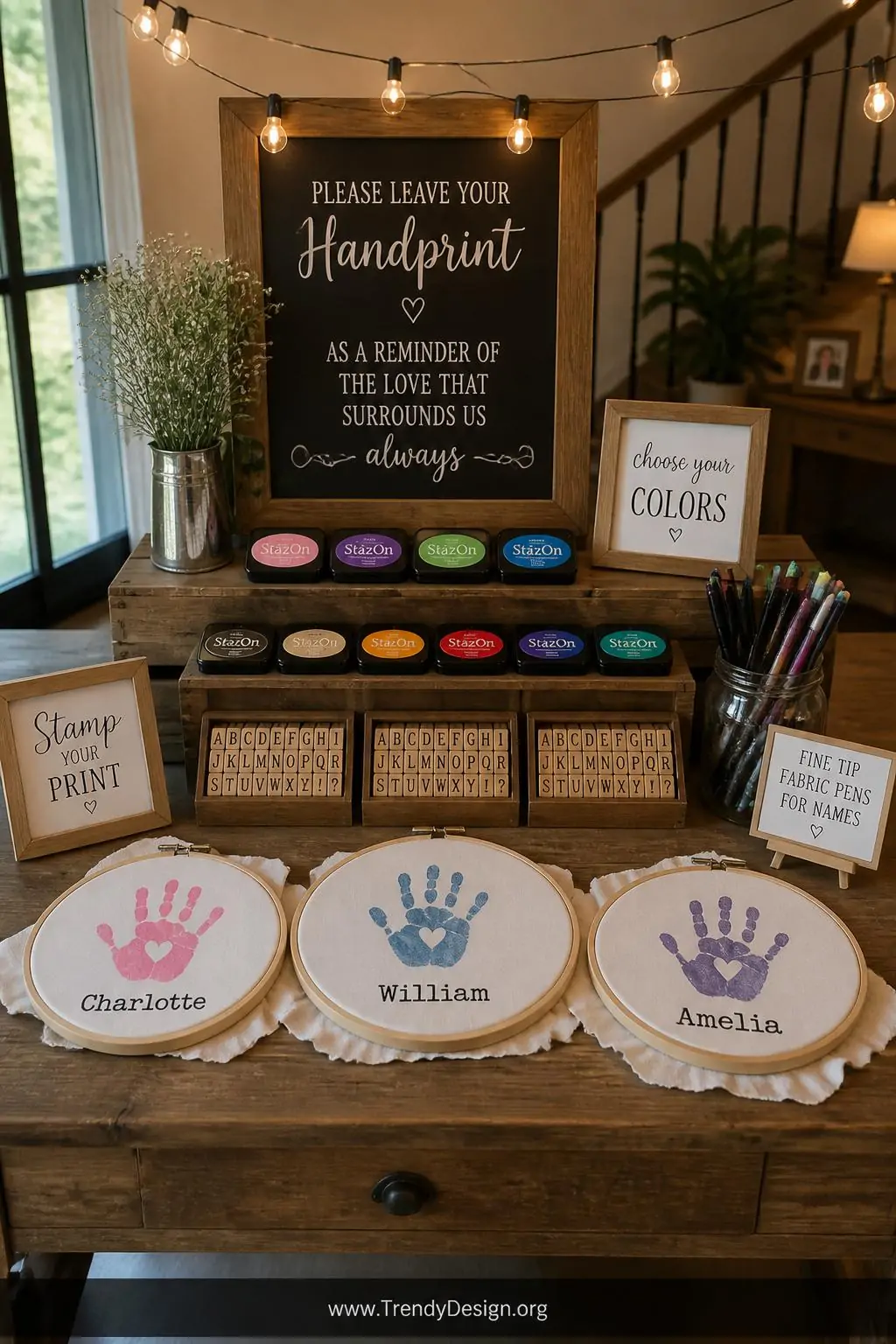 Rustic wooden table vignette with three embroidery hoops holding stretched ivory fabric surrounded by colorful StazOn ink pads, rubber alphabet stamp sets, and fine-tipped fabric pens arranged in a mason jar, warm party string lights glowing overhead, wide shot showing the full interactive craft station setup with stamped handprints and name impressions visible on the taut fabric