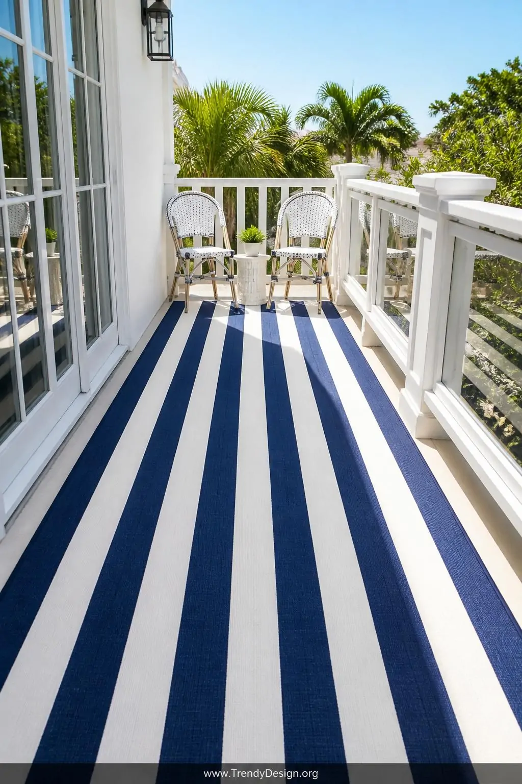 Striped white and navy cabana-style indoor-outdoor rug placed lengthwise down a narrow balcony, wide shot, two white bistro chairs at the far end, the bold parallel stripes visually stretching the compact space, bright midday summer light reflecting off crisp clean furniture and glossy railing.