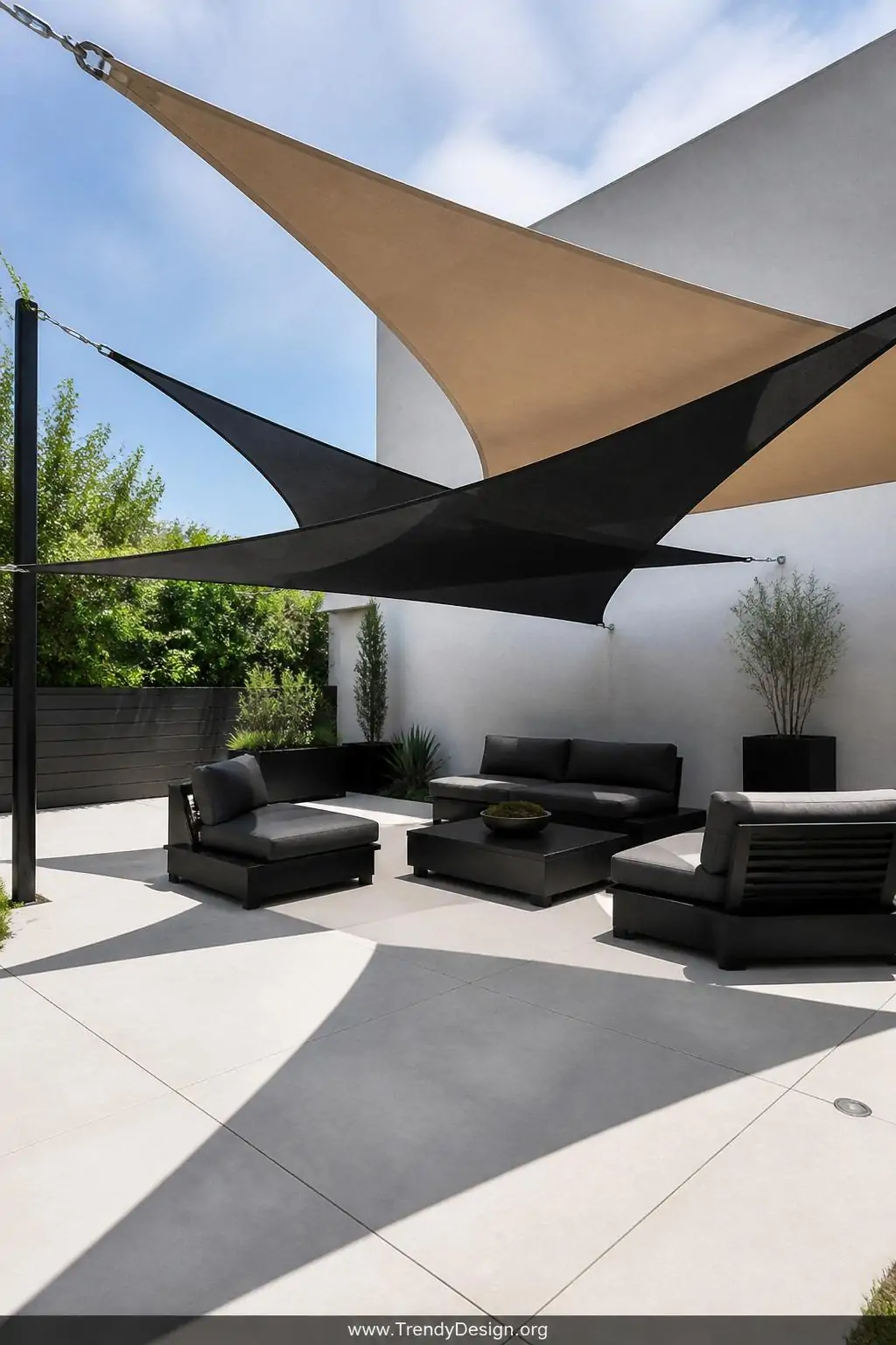 Overlapping triangular shade sails in sandy beige and deep charcoal tensioned at dramatic opposing angles above a minimalist concrete patio with low-profile matte black furniture and geometric planters, wide shot showing stainless steel corner hardware and the bold angular shadow patterns cast across pale pavers below, photorealistic interior design.